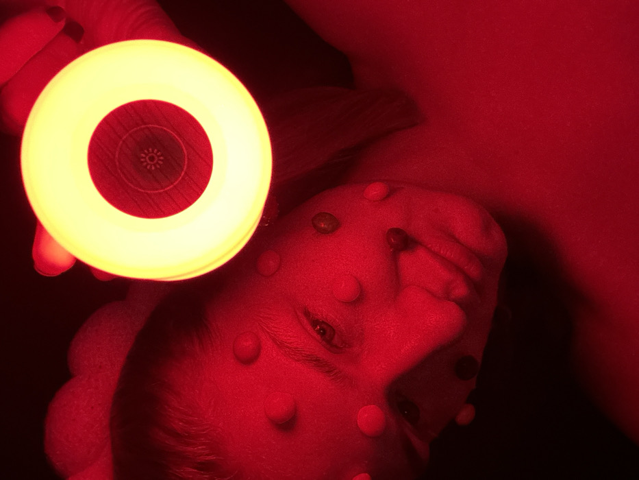 The image, set in Kazan, Russia, presents a close-up, top-down perspective of an individual's face under intense red illumination. The face, likely female, occupies the left and central areas of the frame, with her right eye open and directed generally upwards or towards the viewer. Numerous small, spherical or hemispherical objects, varying in perceived color from red to white and dark, are affixed to the forehead, cheeks, nose, and around the mouth. In the upper-right quadrant, a hand is visible holding a circular light-emitting device. This device projects a bright yellow halo of light surrounding a central red disc, which features a faint radial or starburst pattern in its core. The background is largely undifferentiated and dark, accentuating the subject and the light source.