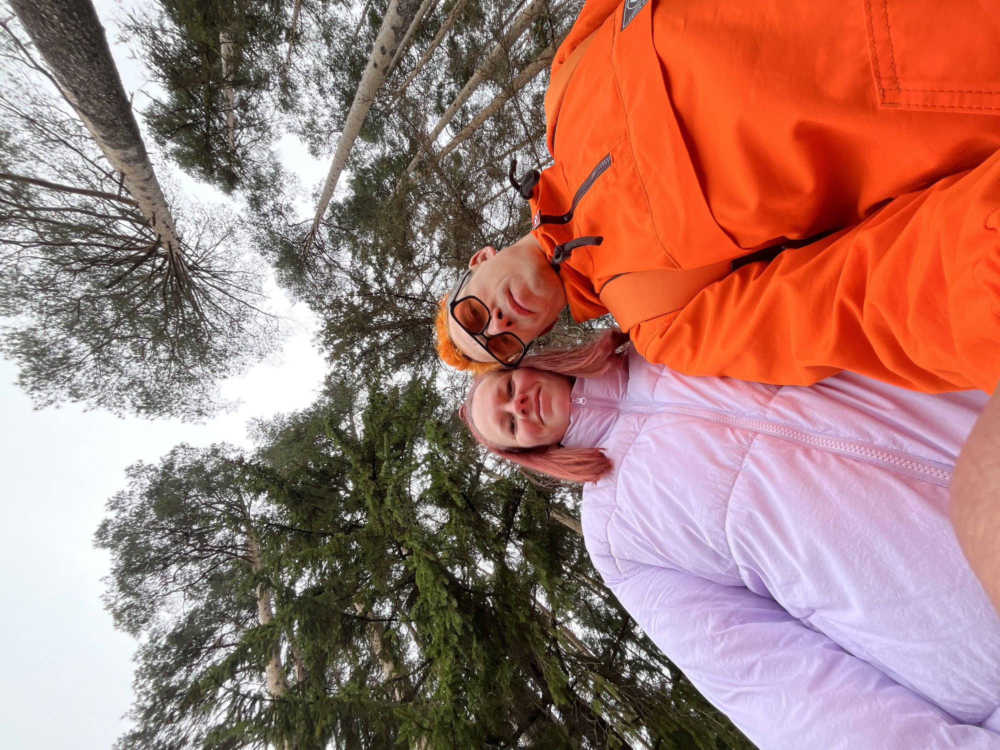 A selfie captured from a low angle featuring two individuals, a person in an orange jacket and a person in a light purple jacket, positioned against a backdrop of tall trees. The location is outdoors, possibly a park or forest in the city of Chaikovsky, Russia. The trees are bare or sparsely covered with leaves, suggesting it might be autumn or winter. The sky is overcast. The person in orange is wearing dark-rimmed sunglasses and has light-colored hair with orange tint, while the person in light purple has pink hair.