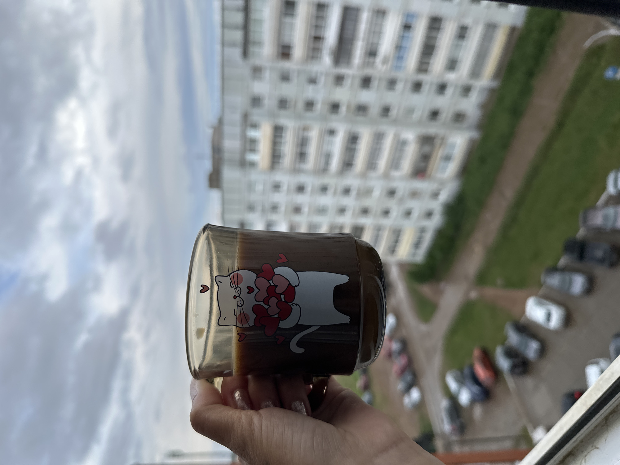 A person's hand holds a glass mug filled with a dark liquid, possibly coffee, featuring a cat graphic holding hearts, against the backdrop of an urban scene. The view from an elevated position, potentially a high-rise apartment, reveals a building complex with numerous windows and a parking lot below populated by several cars. The setting appears to be in Chaikovsky, Russia, based on identifiable architectural style and the arrangement of buildings and vehicles. The sky is overcast, suggesting a cloudy day. The focus is on the mug and the immediate cityscape, providing a glimpse into an everyday moment set against an urban environment.