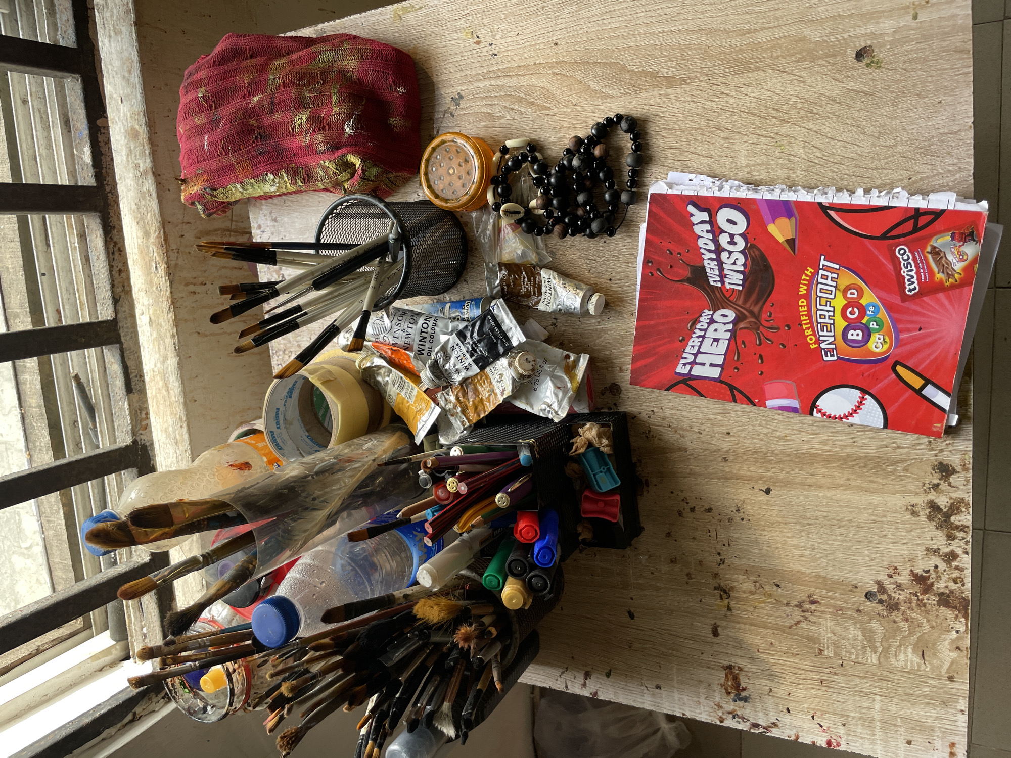 The image depicts an artist's workstation situated on a wooden surface, likely a table or desk, near a window with a metal grate. The scene is filled with art supplies. Prominently displayed are numerous paintbrushes of varying sizes and types, some arranged in clear plastic containers, others in a black mesh holder. Several tubes of oil paint, specifically "Winton Oil Colour" by Winsor & Newton, are scattered among the brushes. Also visible are bottles of water, rolls of tape, colored pencils, markers, and a small orange container with a textured lid. A red, patterned cloth is draped to the right, and a strand of dark beaded jewelry with some shell accents lies near the orange container. In the foreground, a bright red booklet or magazine with "EVERYDAY HERO EVERYDAY TWISCO" prominently displayed is open. The overall environment suggests an active art creation space, with scattered paint marks on the wooden surface. The location is identified as Ado, Nigeria.