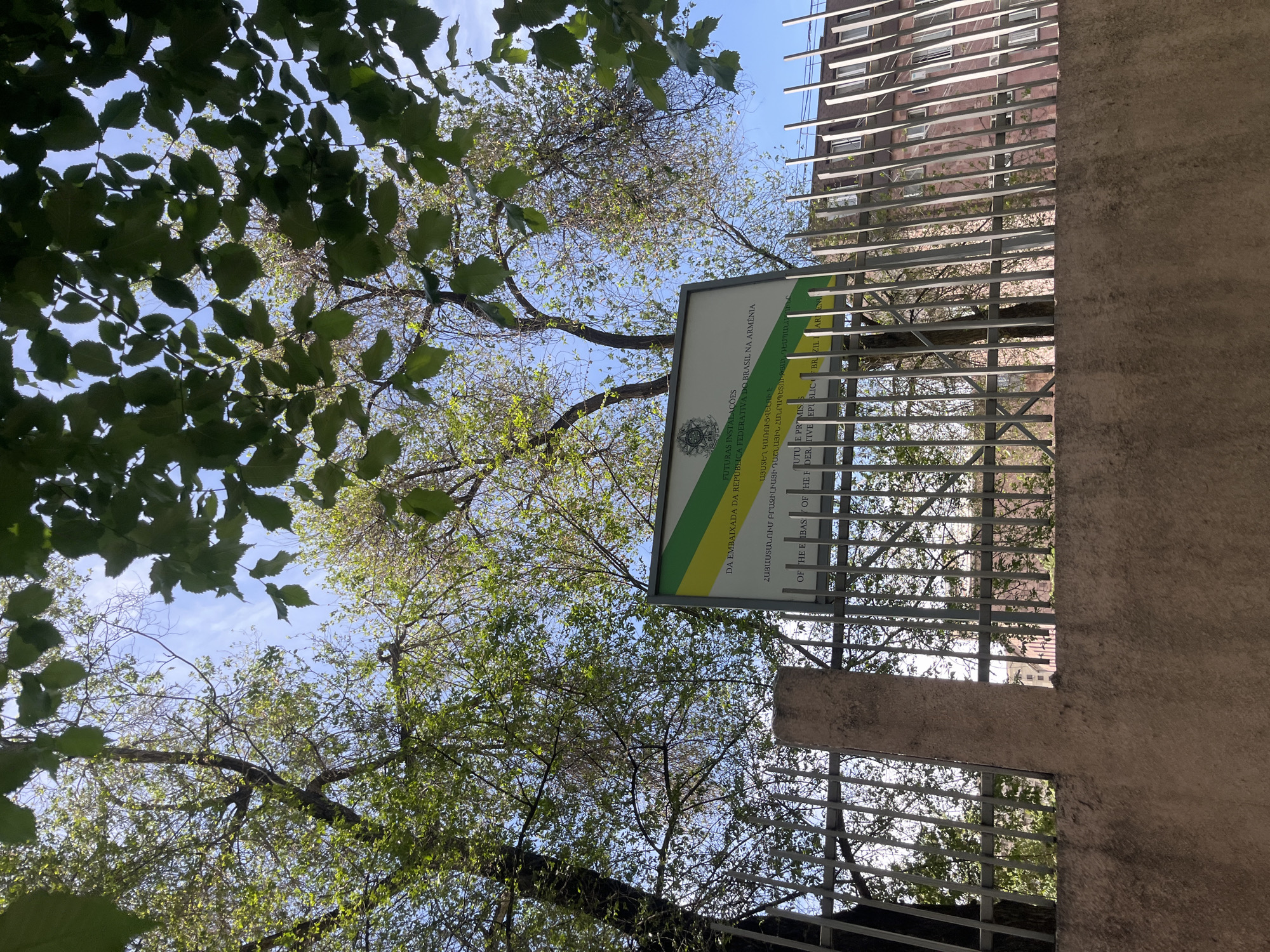 The image shows a sign for the future location of the Embassy of the Federative Republic of Brazil in Armenia. The sign is in Portuguese and Armenian and includes the coat of arms of Brazil. The sign is affixed to a metal fence, and behind it is a building with metal sunshades. Trees with green foliage are visible in the foreground. The sky is blue. The location is Yerevan, Armenia.