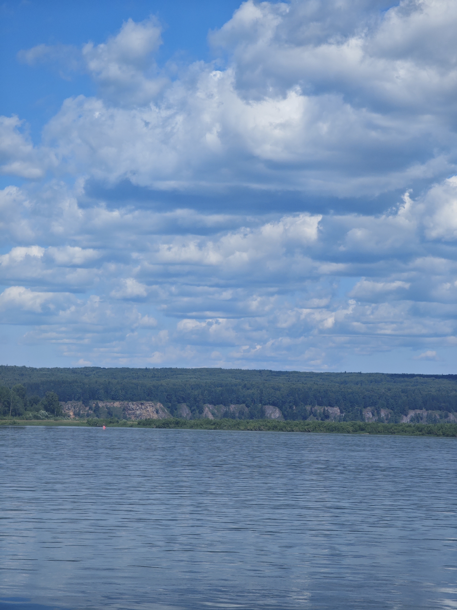 The image depicts a wide, calm river in Strelka, Russia, under a sky filled with dramatic cumulus clouds. The river occupies the lower half of the frame, its surface reflecting the blue sky and exhibiting a pattern of gentle ripples. The opposite bank of the river is lined with a dense, green forest that stretches across the horizon. Above the tree line, a rocky cliff face is visible, interspersed with vegetation. The sky is a vibrant blue, with numerous white and grey clouds creating a textured canopy. A single, small red buoy is visible floating on the water near the distant shore. The overall scene conveys a sense of natural tranquility and vastness.