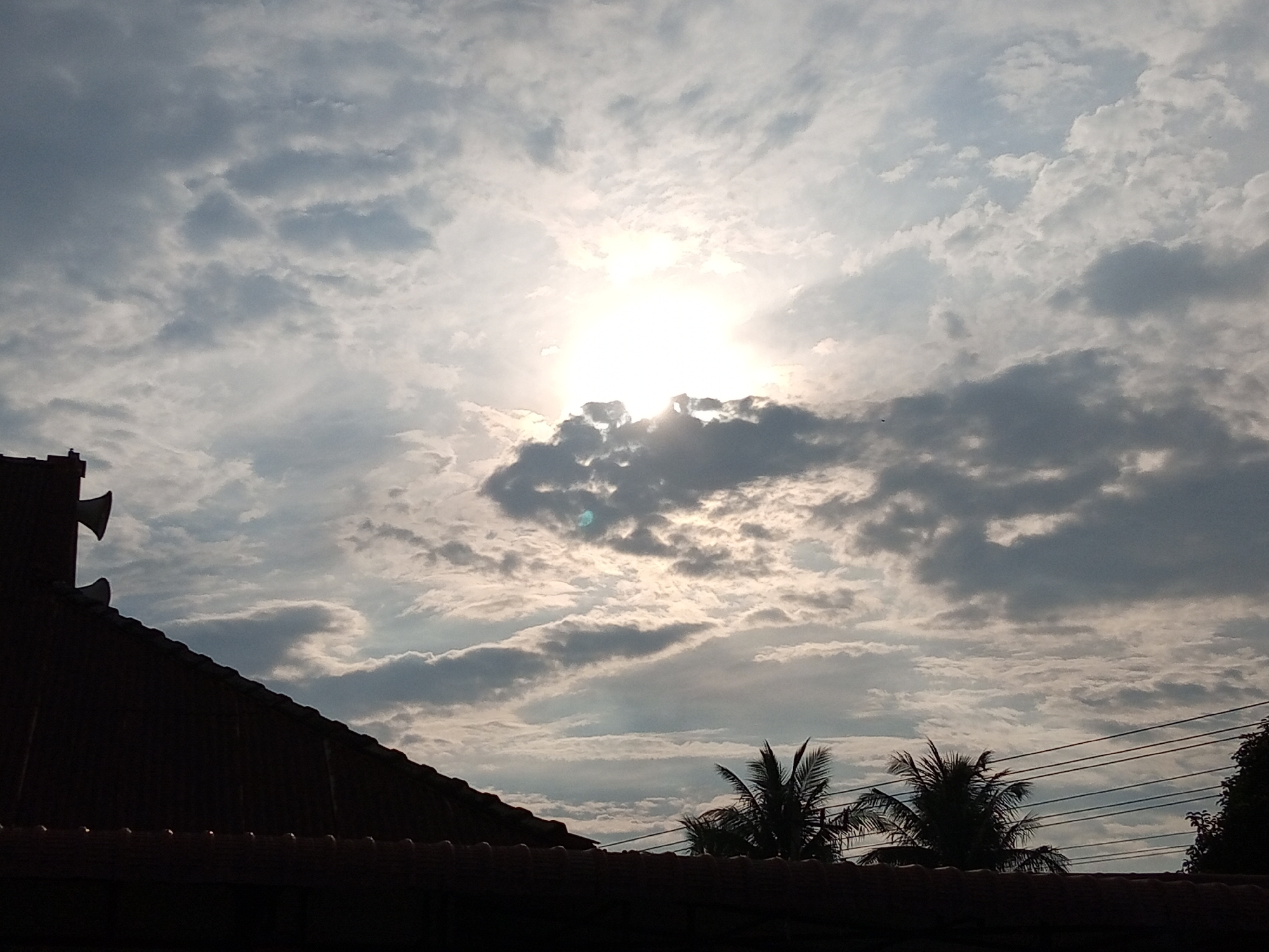 A sky filled with clouds is visible in this image, with the sun shining brightly through them. The sun is positioned slightly off-center to the left. In the foreground, the dark silhouette of a building's roof with tiles is visible, along with a section of a red tiled roof and two conical loudspeakers mounted on a vertical structure. To the right, silhouetted palm trees and electrical wires can be seen against the cloudy sky. The scene appears to be from a city, possibly Cotpuuk, Indonesia, given the presence of tropical vegetation and architectural elements.