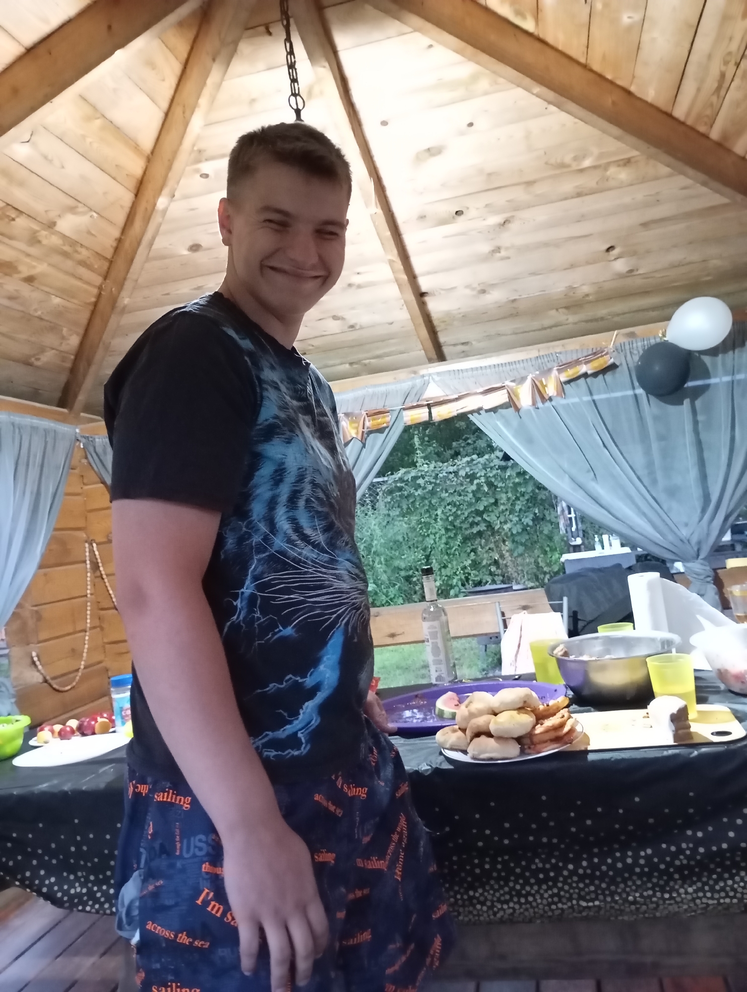 A young Caucasian man with short brown hair smiles widely, looking towards the camera. He is wearing a black t-shirt with a blue and white graphic design of a tiger or cat and blue shorts with a "sailing" motif. The man is standing in an open-air gazebo with a wooden roof and walls. A table is set up in front of him, covered with a black tablecloth adorned with white polka dots. On the table are various food items, including a plate of pastries, a bowl of fruit, and drinks in yellow cups. Party decorations, including a banner and balloons, are visible in the background, suggesting a celebration. The surrounding environment appears to be outdoors, with lush green foliage visible beyond the gazebo. The scene suggests a casual outdoor gathering, possibly a birthday party or a similar event, taking place in Malyi Bukor, Russia.