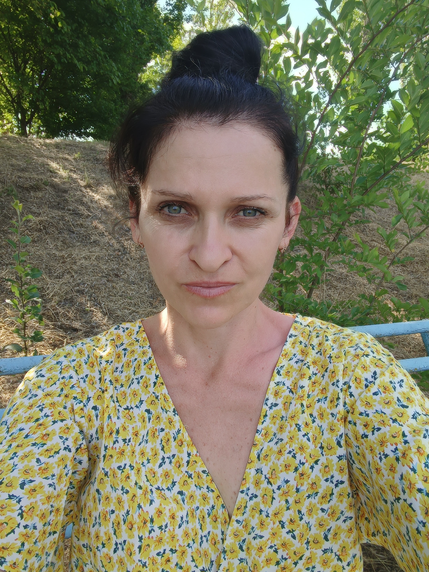 A middle-aged woman with dark hair pulled up in a bun is looking directly at the camera. She is wearing a yellow dress with a floral pattern. The background shows a grassy slope and trees. The scene is outdoors, with natural light. The location is Nezavertaylovka, Moldova.