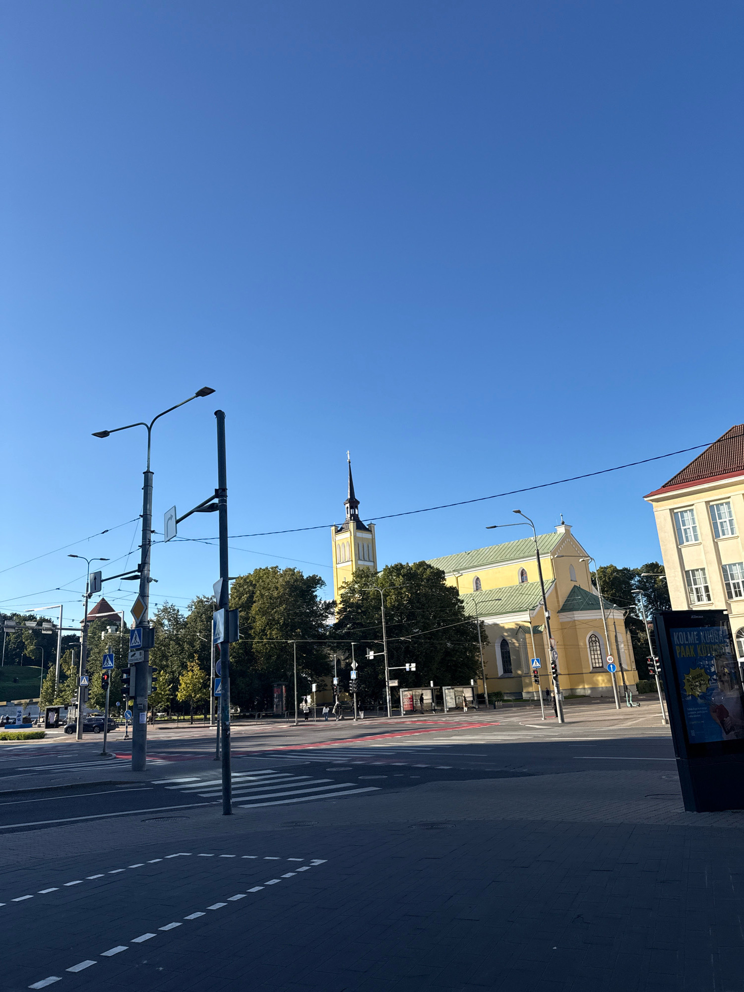 The image shows an intersection in Tallinn, Estonia, with a yellow church with a green roof and a tall spire in the background. To the right of the church is a light-colored building with a red roof. In the foreground, there is a pedestrian crossing with red and white stripes. Several streetlights and traffic signals are visible. A billboard with text in Estonian is located on the right side of the image. The sky is clear and blue. No people are clearly discernible in the image, and no actions or interactions are taking place.