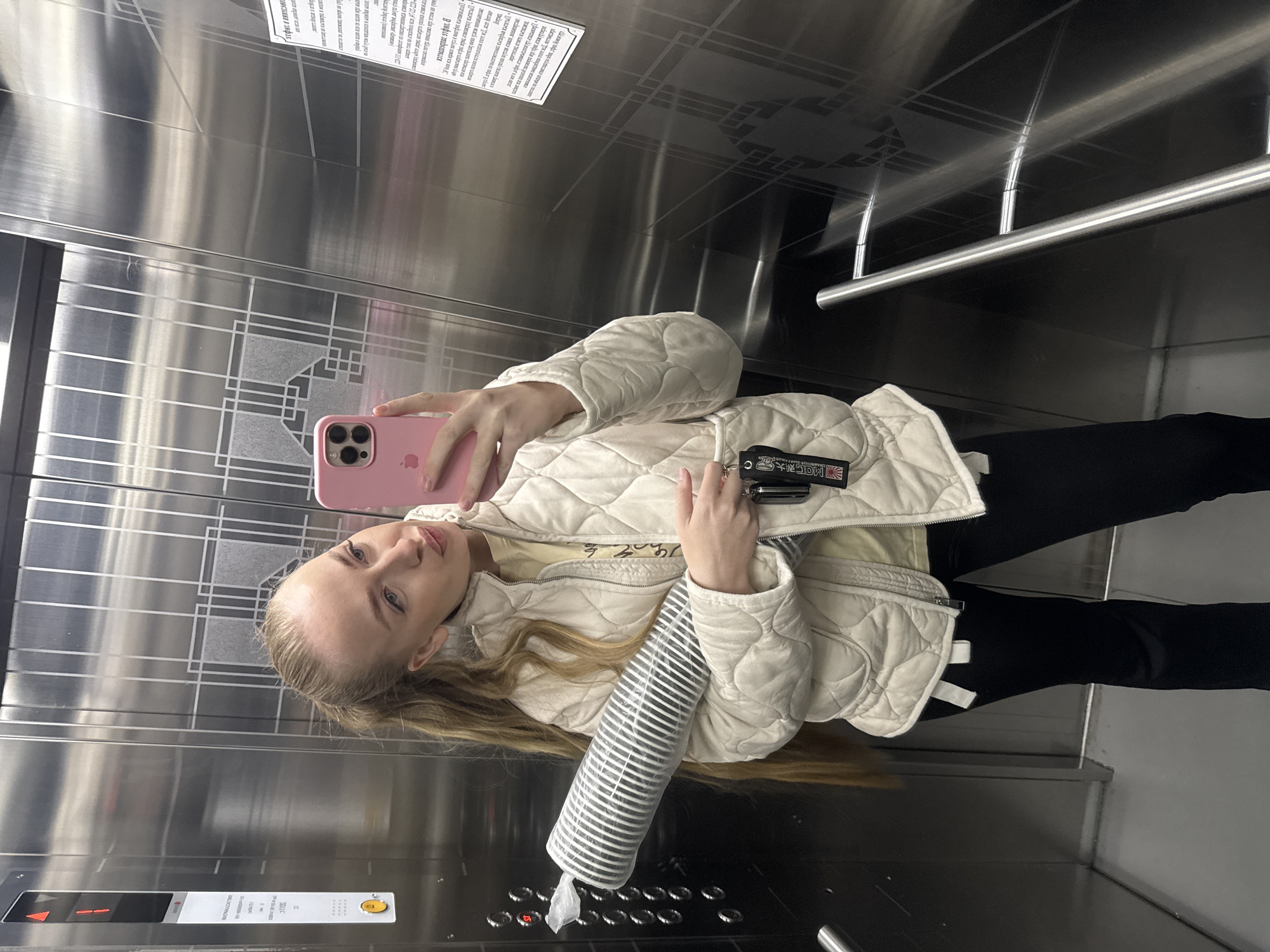 The image shows a young woman in an elevator, likely taking a selfie. She is wearing a light-colored quilted jacket, black pants, and holding a pink iPhone in one hand and a stack of disposable paper cups wrapped in plastic in the other. Her keys are attached to a lanyard or keychain visible near her hand. The elevator has brushed metal walls, an informational sign, and a control panel. The elevator is currently on the first floor. It is impossible to determine the specific location in Petropavlovsk-Kamchatsky from the image.