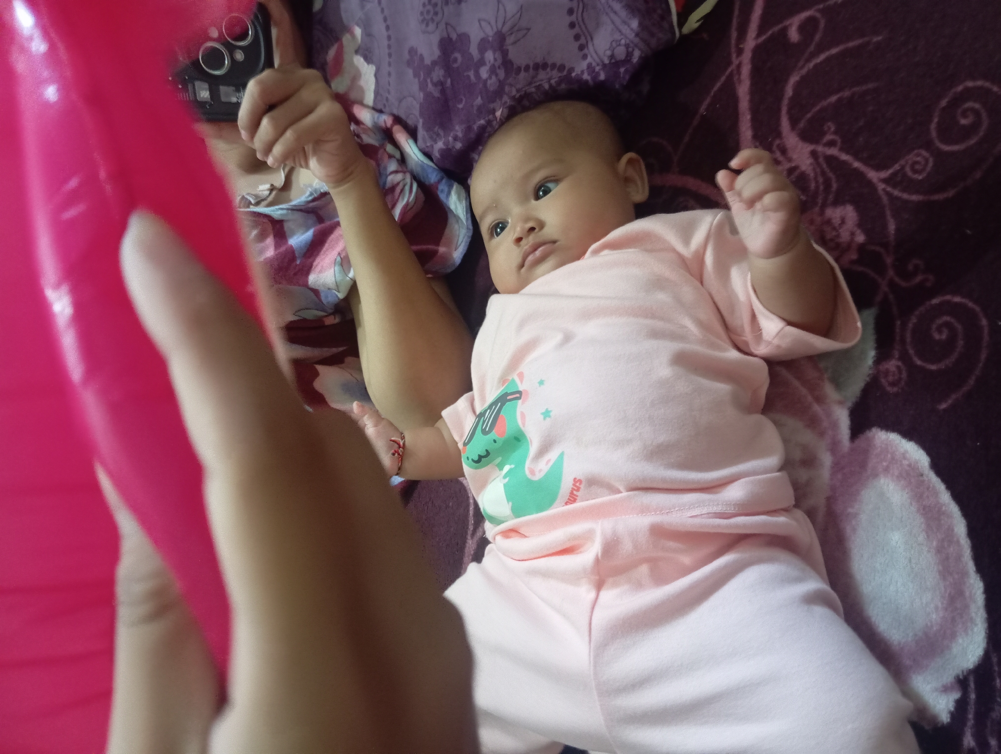 The image shows a close-up of a baby lying on a bed with a pink outfit. The baby is looking up and to the right with a relaxed expression. Part of an adult's arm and hand are visible at the top left, holding a pink object that partially obscures the scene. Another adult's hand is visible in the upper right, holding a smartphone. The background consists of a patterned bedspread. The scene appears to be indoors, likely in a residential setting. No specific location in Rejosari, Indonesia is discernible from the image.