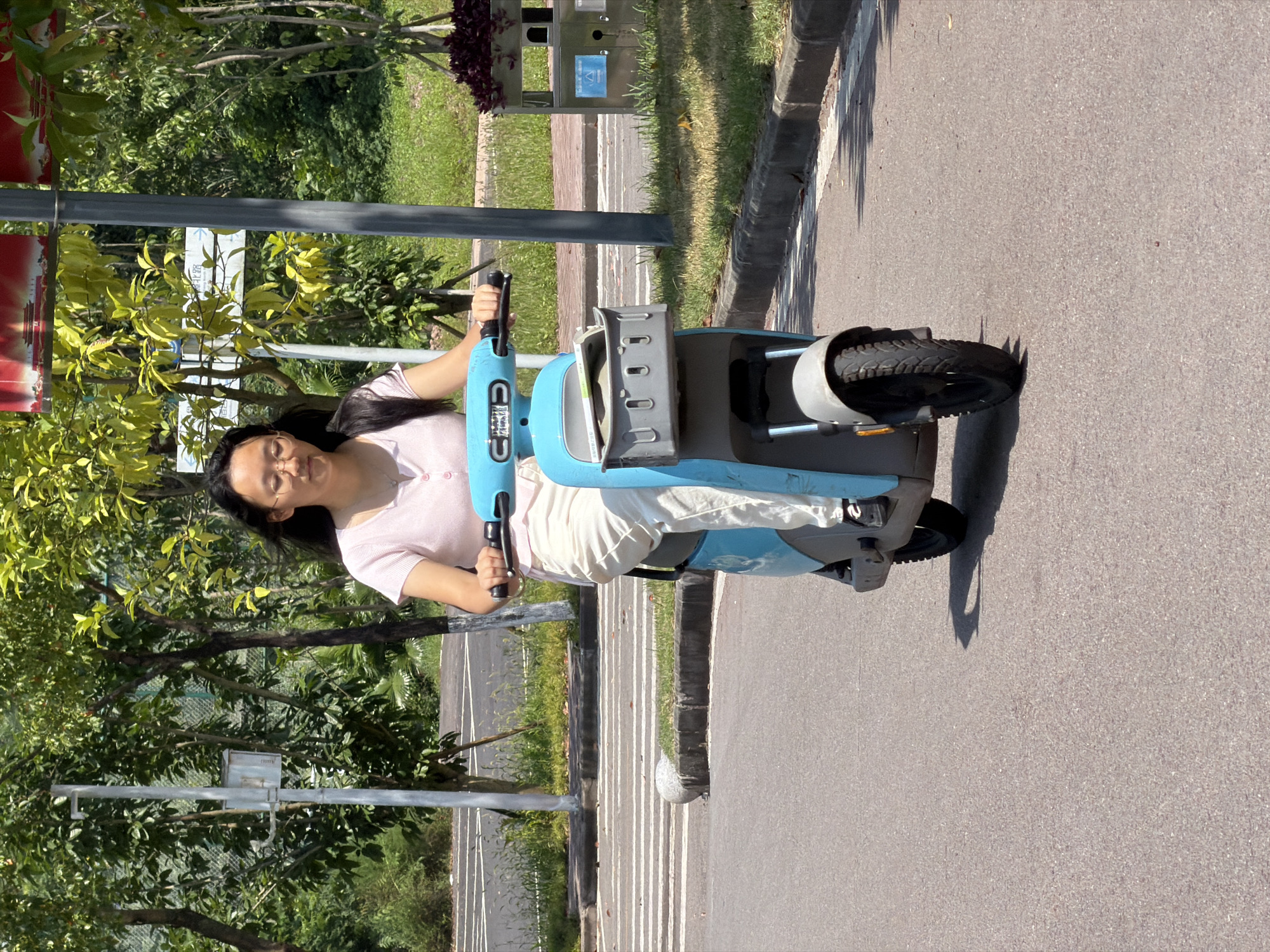 A young Asian woman with glasses and long dark hair rides a blue electric scooter on an asphalt road. She is wearing a light pink short-sleeved shirt and light-colored pants. The scooter has a gray basket attached to the front, containing some items. The scene appears to be outdoors in a park-like setting with trees and greenery visible in the background. A street lamp and a trash can are also present. The road has white markings and a curb. There is a pedestrian crossing in the distance. The lighting suggests it is daytime. This location is identified as Jiangjin District, China.