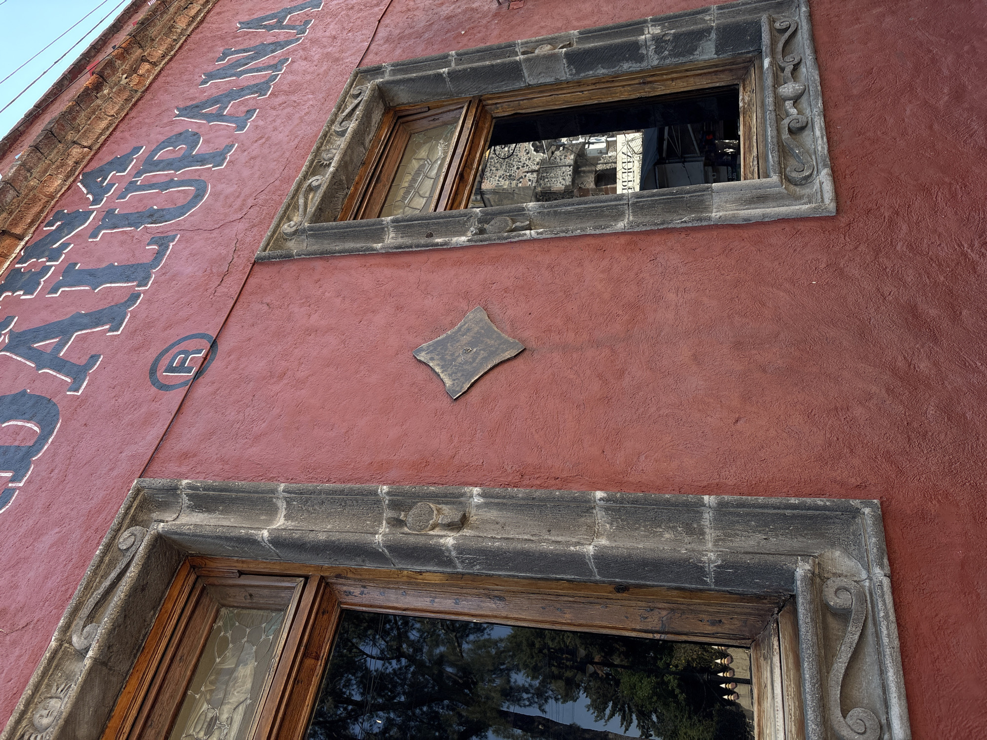 The image presents a detailed view of a building facade in Mexico City, featuring a vibrant reddish-brown stucco wall under clear daylight. Two rectangular windows are the central subjects, each framed by elaborate dark grey stone moldings. These stone frames are intricately carved with decorative scrolls and circular motifs, suggesting traditional Mexican architecture.
The windows themselves have aged wooden frames; the upper window features a pane that is slightly ajar, revealing decorative glasswork, possibly leaded or stained glass. Both windows reflect the surroundings, showing dense green foliage from nearby trees and glimpses of other structures, indicating an urban setting.
Between the two windows, a diamond-shaped decorative plaque, also made of stone, is affixed to the wall. On the upper left side of the facade, large black letters with white outlines are painted vertically, spelling out "ATLATONAPA". Below this text, a registered trademark symbol (®) is visible. The bright blue sky is visible in the top left corner, confirming a sunny day. The overall impression is one of historical charm and rich craftsmanship, typical of Mexico City's older neighborhoods.