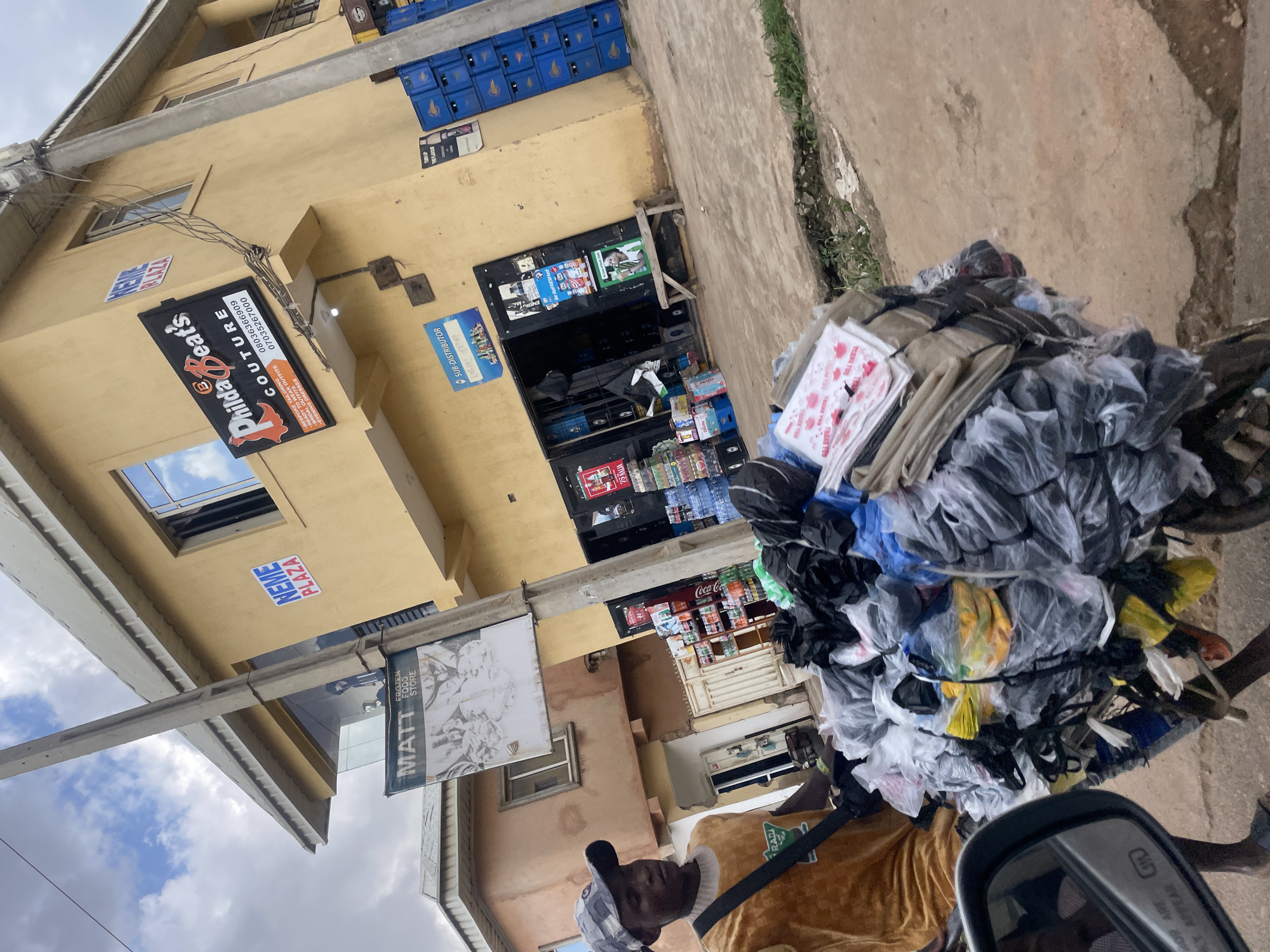 This media file captures a street scene in Gura Top, Nigeria, characterized by a bright yellow building with various storefronts. A person, likely a vendor, is partially visible in the foreground, pushing a cart piled high with goods, predominantly black and clear plastic bags containing clothing or fabrics. The person wears a light brown outfit and a patterned cap.
The building houses several businesses, indicated by signage. One prominent sign reads "PhildaBeat's COUTURE" with contact numbers, advertising tailoring and ready-to-wear outfits. Another sign, partially obscured, says "MATT FROZEN FOOD STORE". Above these, a sign for "NEME PLAZA" is visible. Blue plastic crates are stacked near the top of the building. The storefronts appear to be small shops, with shelves of drinks and other items visible through a glass display.
The scene suggests a bustling commercial area, possibly during daytime, given the lighting. The weather appears overcast, with visible clouds in the sky. The presence of the cart filled with goods and the vendor indicates commercial activity and trade happening on the street. The overall impression is one of everyday commerce in a Nigerian urban or semi-urban environment.