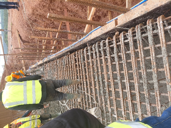 The scene depicts an active construction site in Fdalate, Morocco. Four individuals, identified as construction workers, are present. In the midground, a worker viewed from behind wears a dark shirt, a yellow high-visibility vest, dark work trousers, and mud-splattered green rubber boots, standing on a horizontal grid of steel rebar. To his left, another worker wears a yellow hard hat and a yellow high-visibility vest. A third worker, also viewed from behind, wears a yellow hard hat, an orange high-visibility vest, light-colored pants, and muddy green rubber boots, walking towards the background right. A fourth worker is partially visible in the foreground left, wearing a yellow safety vest and blue jeans.
The primary structure in the image is a densely laid grid of steel reinforcing bars (rebar), partially covered with wet, grey concrete. This rebar grid forms a horizontal structural element. Immediately to the right of this structure is a deep excavation or trench with steep, reddish-brown soil walls. Within this trench, numerous vertical rebar cages are visible, reinforced by horizontal wooden planks and additional rebar. A blue flexible hose runs along the upper edge of the trench, adjacent to the rebar structure. The background consists of undulating reddish-brown earth, consistent with disturbed soil on a construction site, under what appears to be an overcast or hazy sky. The overall environment indicates an ongoing heavy construction project, likely involving concrete pouring for a foundation, slab, or bridge section.