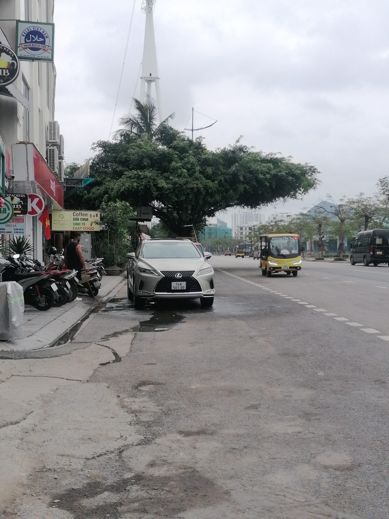 The image shows a street in Bai Chay Ward, Vietnam. A silver Lexus sedan with license plate "14A 501.99" is parked on the side of the road near a row of parked scooters. A man is standing near the scooters holding a long, thin object. The road has visible wear and tear. A yellow electric vehicle and a black van are visible driving on the right side of the road. In the background, buildings and trees are visible under an overcast sky. A tall white tower can be seen in the background. A sign for "Halal Vietnam" is on the building in the background. A sign in front of a business reads "Coffee Sua Chua Sinh To Fast Food".