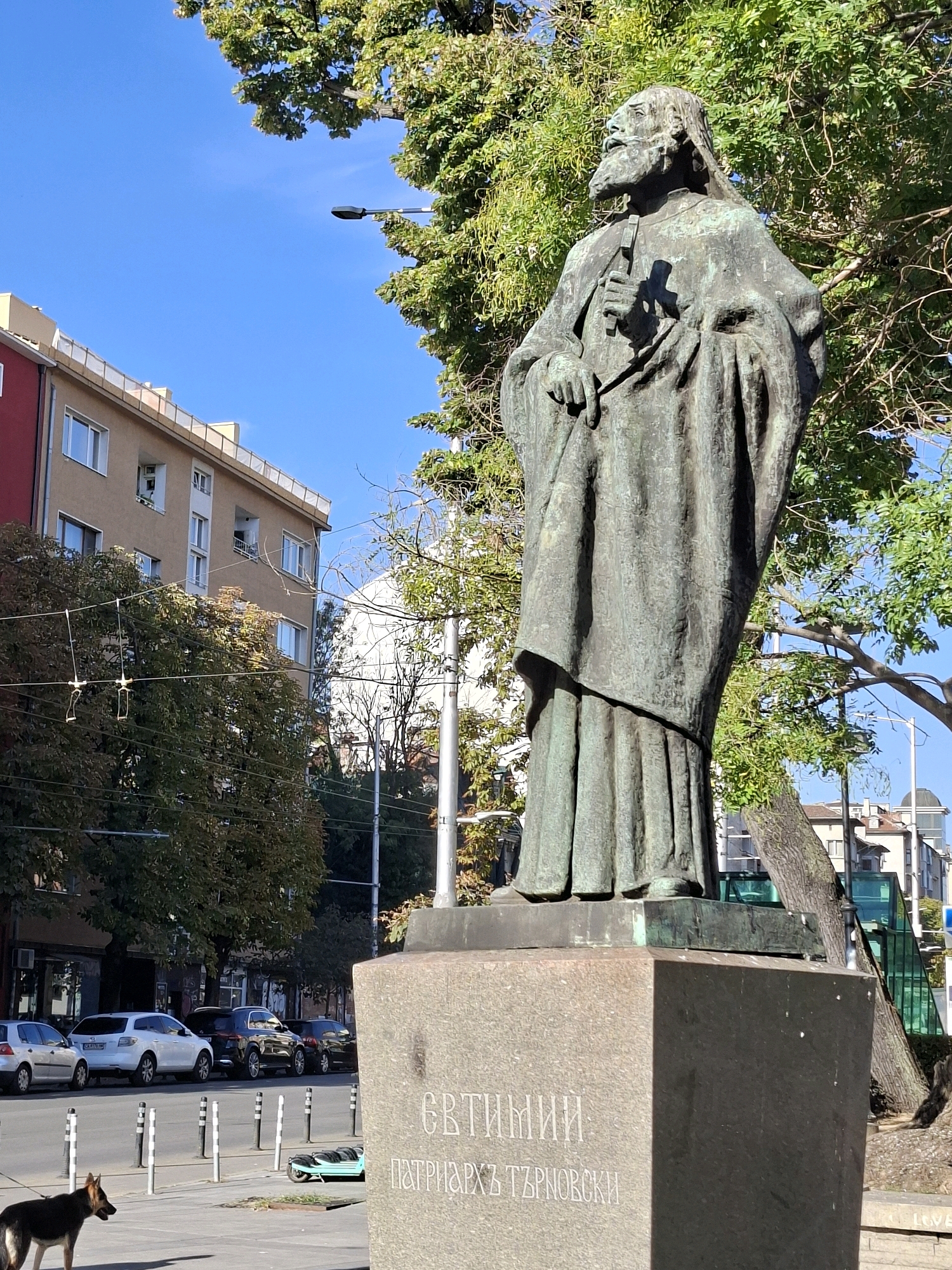An outdoor, eye-level, medium shot shows a bronze statue of a man with a beard wearing a robe and holding a cross in his left hand, standing on a pedestal inscribed with Cyrillic text. The statue is located in a city and is surrounded by trees with green foliage. In the background, there are buildings, a street with parked cars, a lamppost, and a clear blue sky. A German Shepherd dog is visible in the lower left corner of the frame, walking on a sidewalk. The statue is made of bronze and has a weathered patina. The pedestal is made of stone and has carved lettering in Cyrillic. The buildings in the background are modern and multi-story, with balconies and windows. The street is paved and lined with bollards. The dog is black and tan and is wearing a leash.