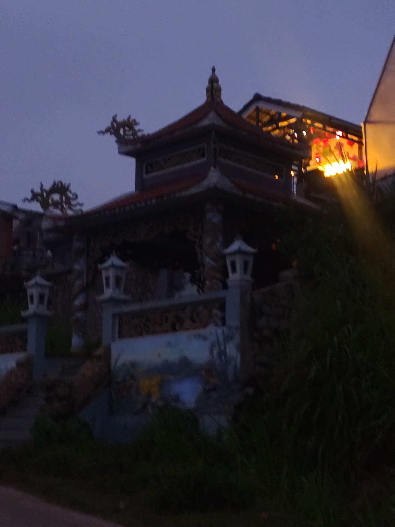 The image depicts a structure with Asian architectural elements, possibly a temple or pavilion, in what appears to be a twilight setting. The structure has multiple tiers, ornate roof decorations including dragon-like figures, and is illuminated by internal lighting. Decorative lanterns are visible in the structure. Below the main building, there is a retaining wall with a mural or painting. In the foreground, vegetation partially obscures the view. A paved area, likely a street, is at the very bottom of the frame. The low lighting suggests the photo was taken at dusk or dawn.