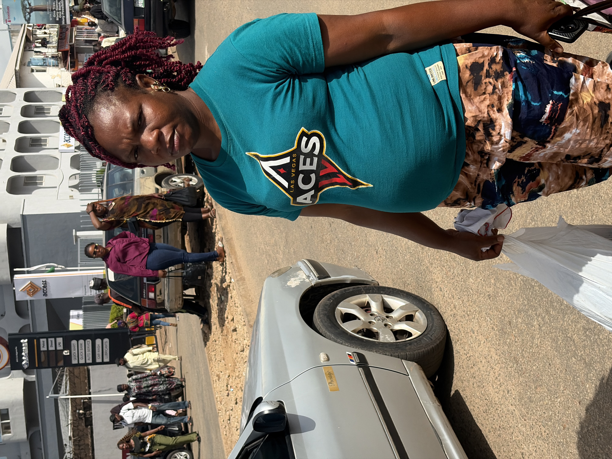 This image depicts a street scene in Jos, Nigeria, bathed in bright sunlight, suggesting a warm, daytime setting. The primary subject is a woman with deep brown skin and dark, braided hair adorned with burgundy highlights. She is wearing a teal t-shirt with a "Las Vegas Aces" logo and a patterned skirt in earthy tones. Her expression is serious, and she is holding a white plastic bag in her right hand.
In the background, several other individuals are visible, some walking along the road, others near vehicles. A silver car is prominent in the foreground, its wheel turned towards the viewer. A gas station with "Access" signage and fuel price information ("Patrol", "LPG", "Diesel") is also clearly visible, indicating a commercial area. Other text includes "Prestige" on the car and "Unlimited Quality" on a tag on the woman's shirt, and what appears to be a business name "bacbab" possibly related to "One Tree, Many Names" and "One Bank, Many Possibilities". The overall atmosphere seems to be that of a bustling urban environment.