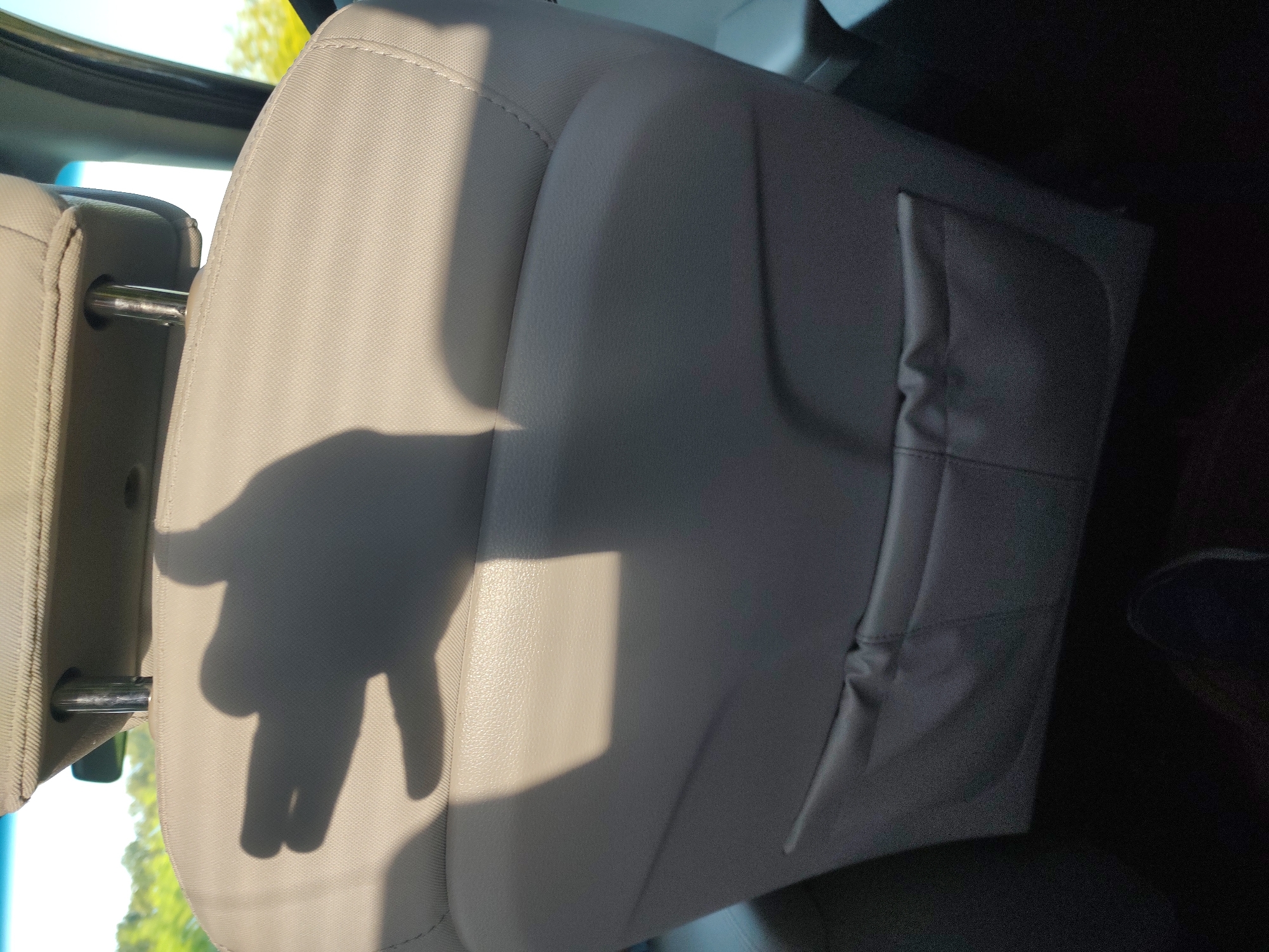 The image shows the interior of a vehicle, specifically the back seat. The seat is upholstered in a light-colored material, likely leather or vinyl, and features a textured pattern and stitched seams. A shadow of a hand making a "peace sign" is cast on the seat, suggesting someone is inside the vehicle and interacting with the light source. The background shows a glimpse of the outdoors, with sunlight filtering through trees, indicating it's daytime. The setting appears to be a sunny day, possibly in a rural or suburban area given the greenery. The overall mood is casual and perhaps playful due to the hand gesture. There is no visible text in the image. The location context provided is Smolnitsa, Bulgaria.