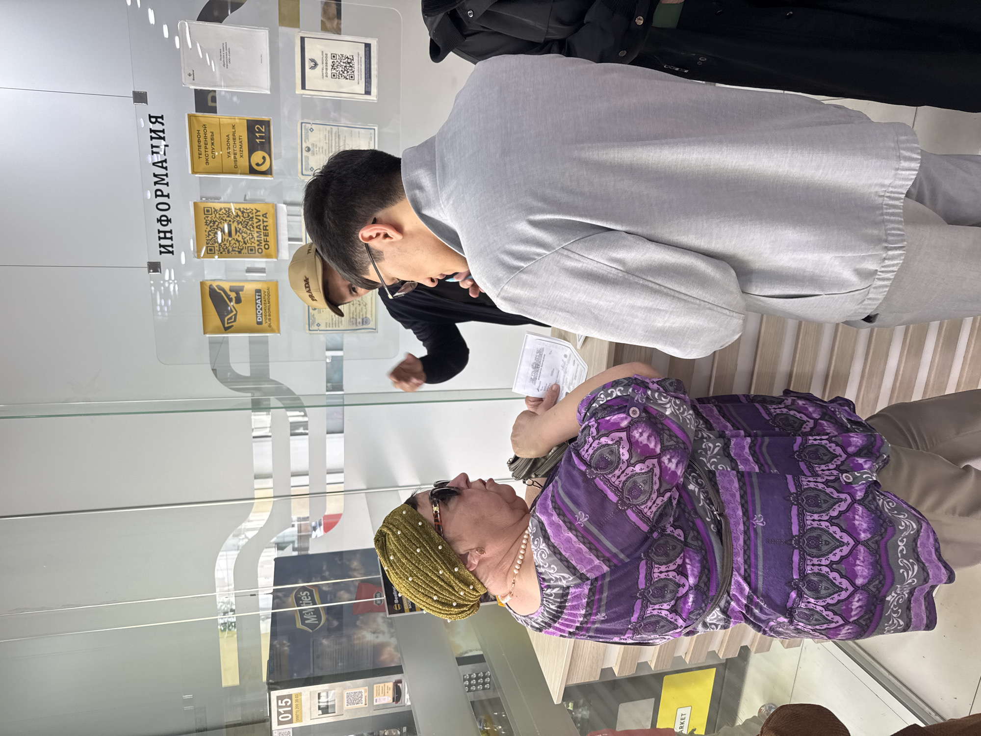 The image shows three individuals in an indoor setting that appears to be a public space in Tashkent, Uzbekistan, likely an office or customer service area. A woman wearing a patterned purple top, a gold head covering, sunglasses, and a pearl necklace is engaged with a young man in a light gray suit. The man is looking down, presumably at paperwork or some object held by the woman. Another man wearing a Prada cap and a dark jacket is partially visible behind the man in the suit. In the background, there is a glass partition with information displayed in both English and a Cyrillic script. This includes notices related to video surveillance ("DIQQAT! VIDEOKUZATUV OLIB BORILMOQDA") and contact information for emergency services ("ТЕЛЕФОН ЭКСТРЕННОЙ СЛУЖБЫ - YAGONA DISPETCHERLIK XIZMATI - 112"). Other elements include signs with QR codes and the number "015" visible near a shelf containing confectionery. The people seem to be engaged in a transaction or information exchange.