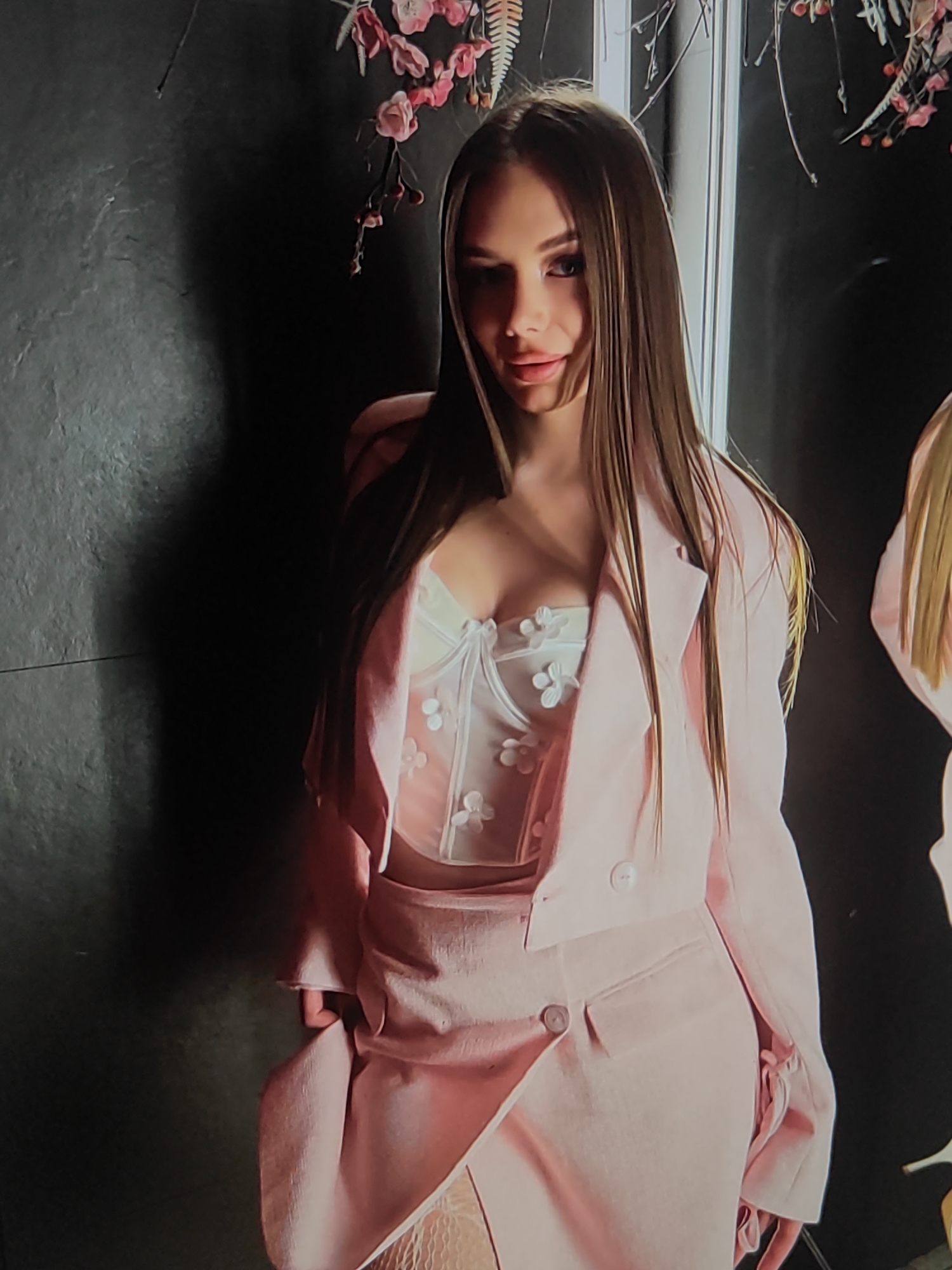 The media file shows a young woman posing against a dark, textured wall. She has long, straight, light brown hair and is wearing a pink blazer over a white corset-style top adorned with floral appliqués. The top appears to be sheer in parts, revealing some of her chest. She is also wearing a matching pink skirt with a similar asymmetrical hem and button detailing. The overall style of her outfit is fashionable and suggestive.
Behind her, a branch with pink flowers and some greenery hangs down, adding a touch of nature to the otherwise stark setting. To her right, another person, mostly out of frame, is visible, also wearing a similar light-colored outfit. The lighting in the image is soft, casting shadows on the wall behind the woman, suggesting an indoor setting, possibly a studio or a well-decorated room.
The woman's expression is alluring, with her lips slightly parted and her gaze directed towards the camera. Her posture is confident and poised. The combination of her attire and the styling suggests a fashion shoot or a similar event. There is no visible text in the image. The location context provided is Fergana City, Uzbekistan, but there are no specific visual cues within the image itself to confirm this location. The time of day and weather are not discernible from the image.