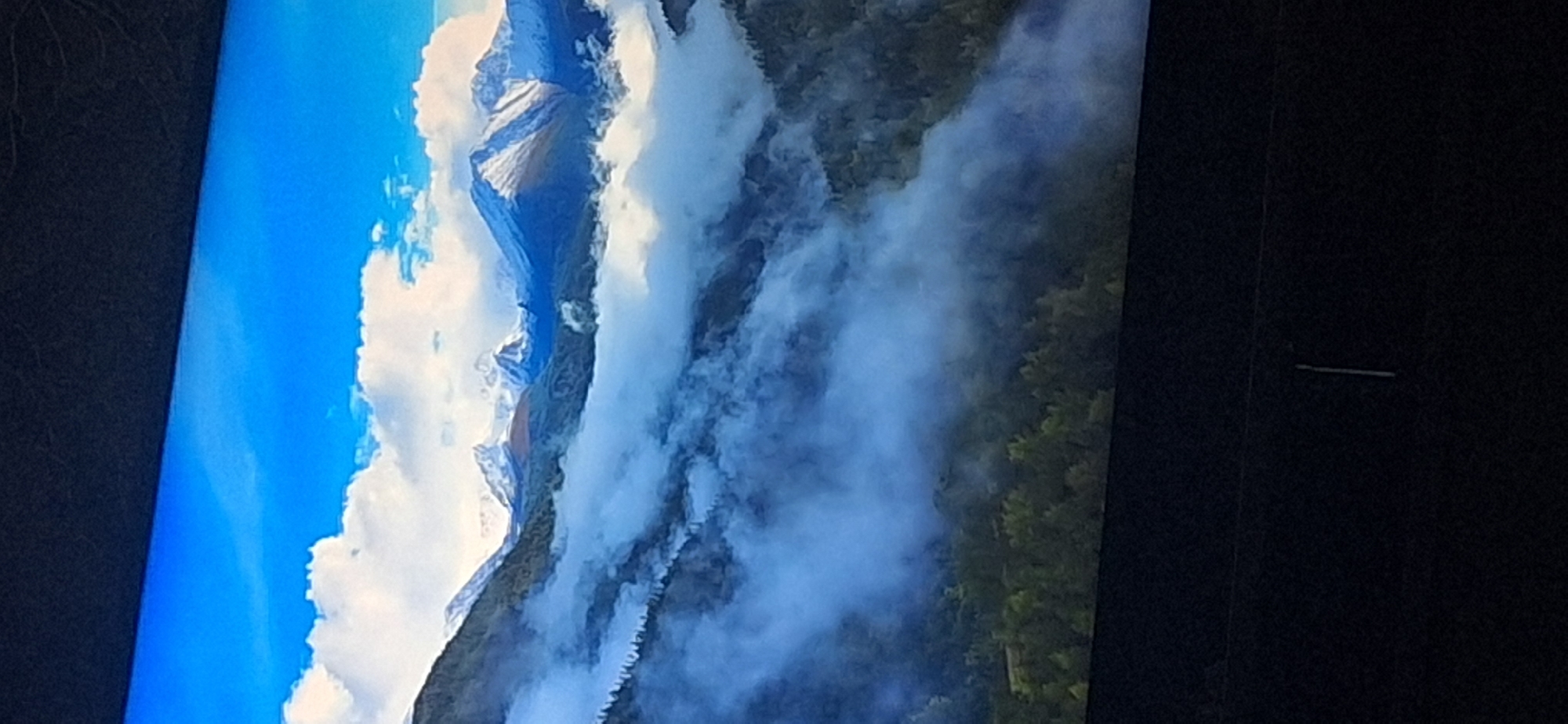 The image displays a sweeping, panoramic view of a mountain range partially obscured by swirling clouds and fog. The peaks of the mountains are capped with snow, contrasting with the darker, forested slopes below. The sky above is a vibrant blue with scattered white clouds. The scene evokes a sense of natural grandeur and is devoid of any human presence or urban elements. The image's orientation is vertical. There is no information within the image to suggest a location in Bila Tserkva, Ukraine.