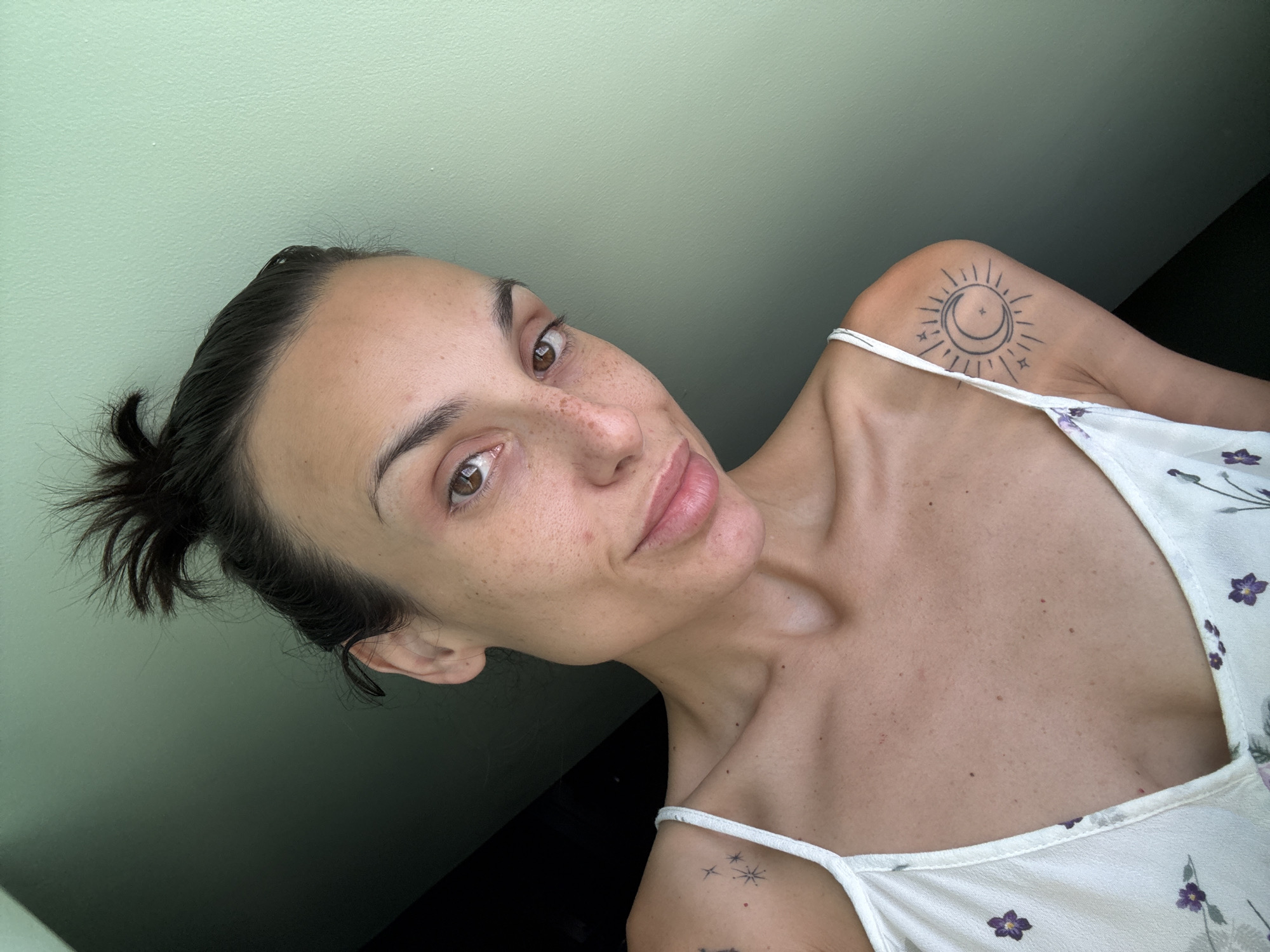 A woman with dark hair pulled up in a messy bun, no makeup, and freckles on her face, looks at the camera. She is wearing a white tank top with a floral pattern. There are two tattoos visible: one of stars on her left shoulder and a sun and moon design on her right shoulder. The background is a wall painted a muted green. The scene is indoors, likely in a room with natural light coming from the side. There is no visible indication of the city of Pleven, Bulgaria.