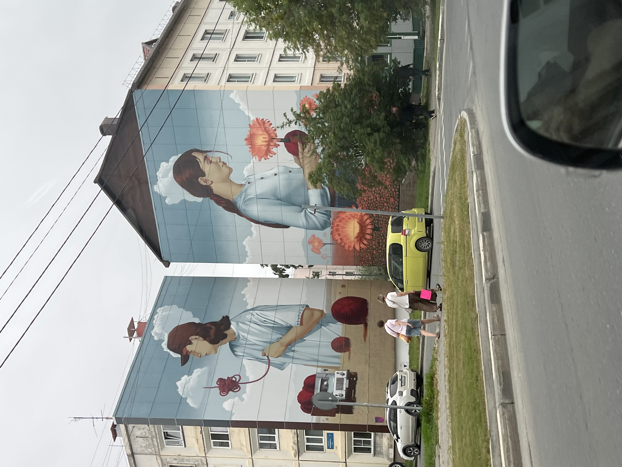 Two murals of young women are painted on the sides of adjacent apartment buildings in Yuzhno-Sakhalinsk, Russia. The mural on the left depicts a woman wearing a baseball cap, holding a red ball of yarn and a strand of yarn. A truck loaded with red yarn balls is visible on the ground in the mural, along with two large red yarn balls on the ground. The mural on the right shows a woman holding a large orange flower and a red ball. The background of both murals features a light blue sky with clouds and stylized orange flowers. In front of the buildings, two women walk along a sidewalk. A white SUV is parked on the street, and a yellow hatchback is driving away. Several trees are visible between the buildings and the street.