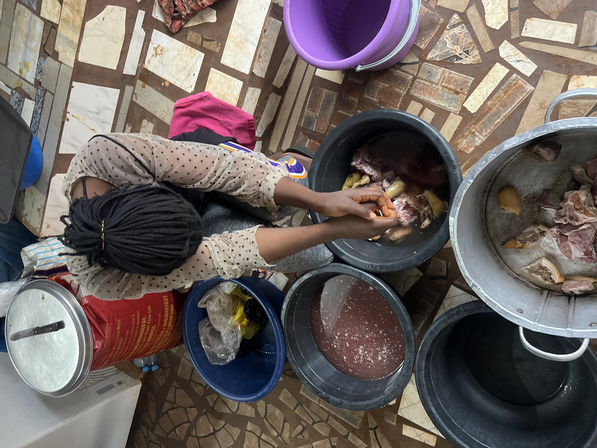 The image captures an overhead view of a person with dark braided hair engaged in food preparation in what appears to be an outdoor or semi-outdoor setting in Nkpor, Nigeria. The ground is tiled with a mosaic of brown and beige squares.
The central figure, viewed from the back and side, wears a sheer, polka-dotted blouse over a darker garment. They are actively washing pieces of meat submerged in a large, dark basin filled with reddish-brown liquid. Their hands are immersed in the basin, manipulating the meat.
Surrounding this basin are several other containers. To the left, a smaller blue basin holds plastic bags, one containing yellow liquid, possibly oil. Below that, a large black basin is empty. To the right of the main basin is another large black basin, also containing water and some meat. Further to the right, a bright purple basin holds clear water. In the foreground, a large metal pot contains more raw meat.
Behind the figure, a large red sack, labeled "Parboiled Rice," is partially visible, topped with a silver lid. Various other items, including plastic bottles and a checkered cloth, are scattered around the area, contributing to a utilitarian and domestic atmosphere. The overall scene depicts a hands-on, traditional approach to cooking.