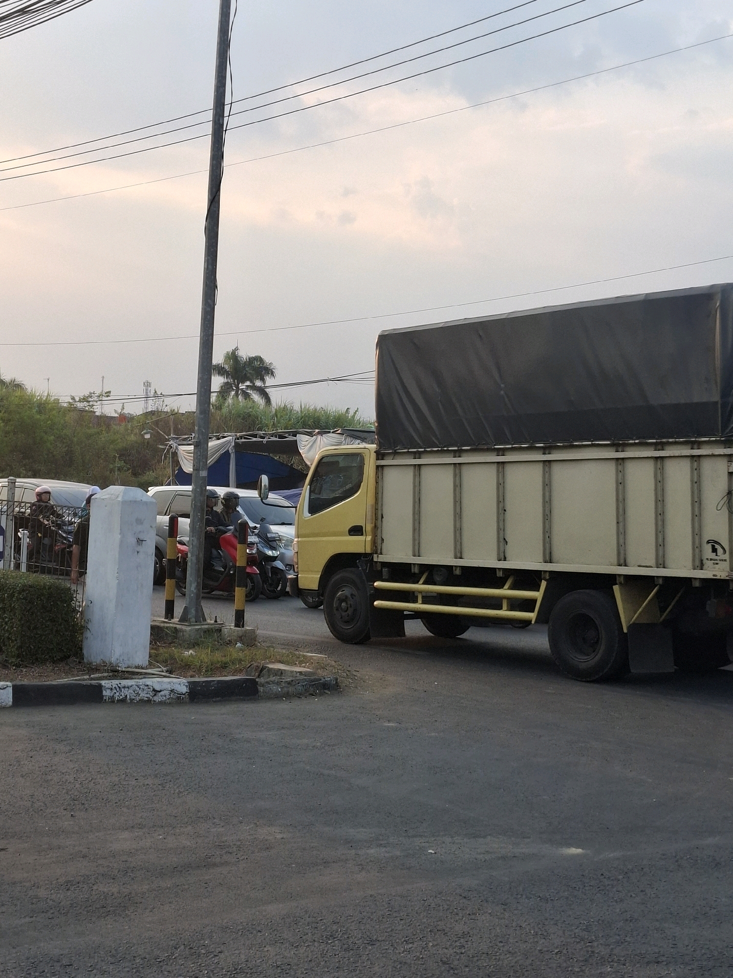 In Karangtengah, Indonesia, a yellow truck with a black tarped cargo bed is parked on a paved road, facing right. The truck has a beige cargo box with vertical ribbed panels and yellow protective bars on the front. To the left of the truck, a line of vehicles is visible, including a red motorcycle with two riders wearing helmets and a white car. A light-colored concrete pillar with a black and yellow striped base stands in the foreground, near a patch of overgrown grass and bushes. A metal pole, likely a utility pole, is positioned behind the pillar, with electrical wires running overhead. The sky above is overcast with a soft, hazy light.