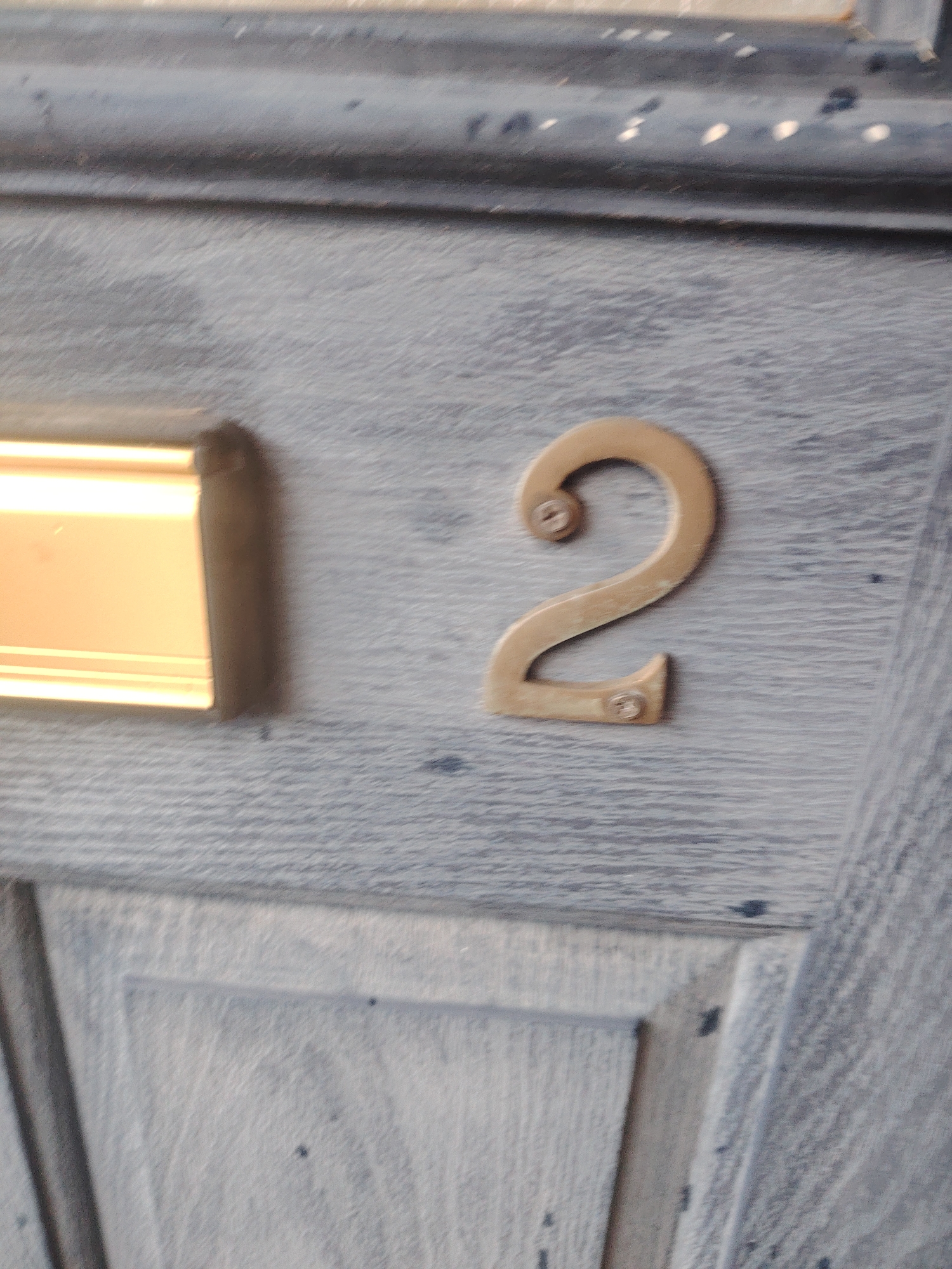 The image shows a close-up view of a weathered, light blue wooden door. The door has a horizontal groove running across its width near the top. To the left of center, there is a rectangular brass or gold-colored mail slot with a beveled edge. To the right of the mail slot, a brass number "2" is affixed to the door. The number "2" is mounted with two small screws, one near the top curve and one at the bottom tail. The overall impression is of an aged exterior door, likely in a residential or commercial property. The setting is not specifically identifiable as being in Huntingdonshire, United Kingdom, based solely on the visual information provided in the image.