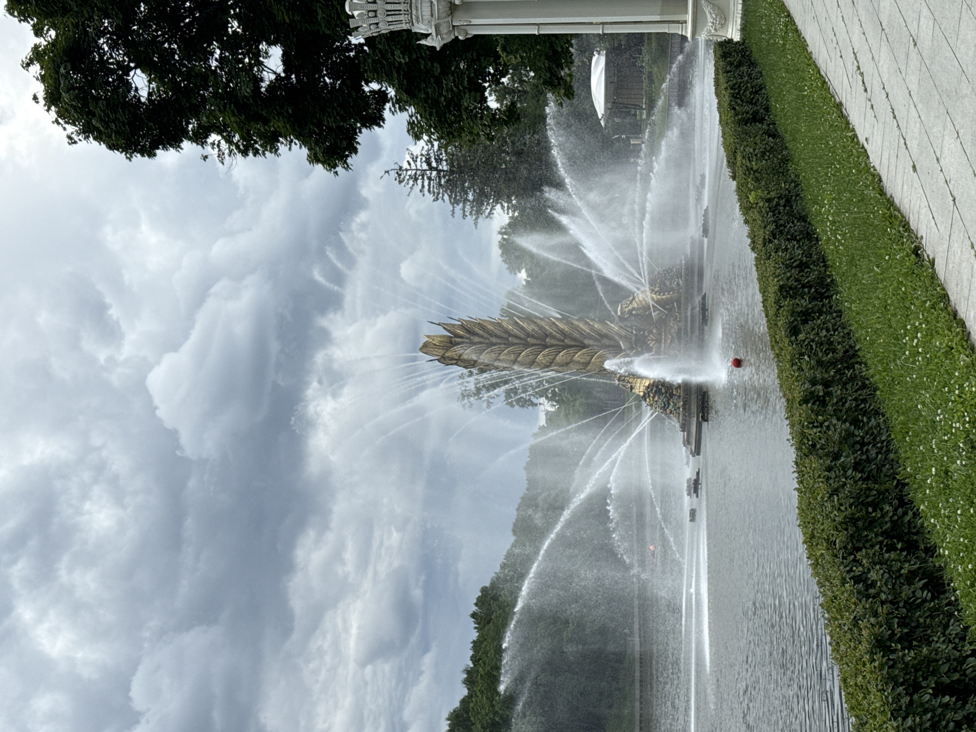 This media file captures a grand fountain in operation, with water spraying upwards and outwards in a dynamic display. The fountain's central sculpture appears to be ornate and possibly gilded, resembling a large ear of wheat or a similar botanical element, supported by more elaborate gold-colored figures. Water streams arc gracefully from the sculpture, creating a misty veil that partially obscures the background.
The scene is set outdoors, likely in a park or formal garden. To the right of the fountain, a meticulously manicured hedge runs parallel to a paved walkway or patio, suggesting a well-maintained public space. Beyond the hedge, a vast expanse of lush green grass stretches out, dotted with small white flowers. In the background, mature trees with dense foliage frame the fountain, and a white architectural structure, possibly part of a building or monument, is visible on the left. The sky is overcast with heavy, dramatic clouds, indicating a day with potentially changing weather. A single red object, perhaps a ball or a marker, is visible in the water near the hedge. The overall impression is one of a majestic and lively water feature in a serene, landscaped environment. No people are clearly visible, and no text is present in the image. The lighting suggests it could be daytime, but the overcast sky might mute the intensity of the light.