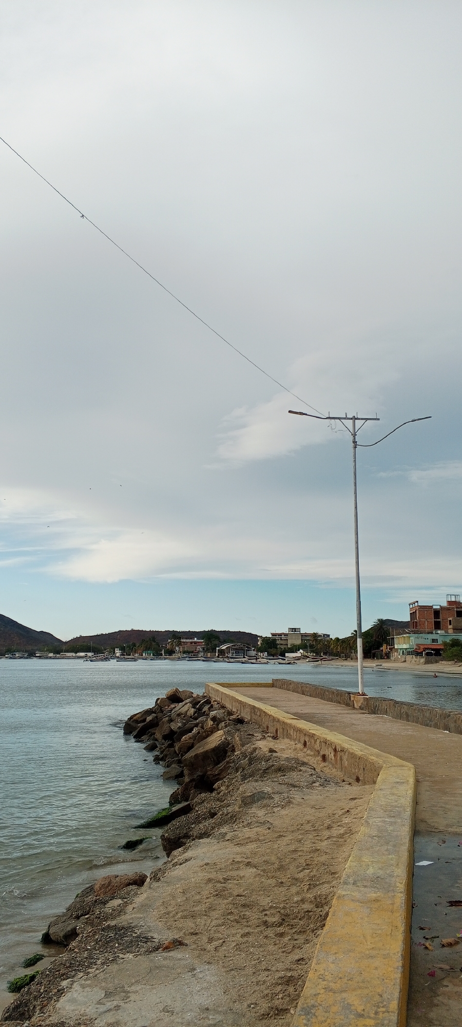 The image captures a waterfront scene in Juan Griego, Venezuela, featuring a concrete pier extending into the calm, grey-blue water of a bay. The pier's edge is lined with large, rough stones, some partially submerged, acting as a breakwater. A sandy embankment slopes down from the pier on the right side, showing scattered debris and some green vegetation clinging to the rocks.
In the distance, the coastline is dotted with buildings of various sizes and architectural styles, including some multi-story structures that appear to be hotels or apartment complexes. A low, verdant hill rises behind the buildings on the left. Numerous small boats are anchored in the bay, suggesting a fishing or recreational harbor.
A tall, grey utility pole with two streetlights stands prominently on the pier, casting a shadow. A thin wire stretches diagonally across the sky from the upper left corner of the frame towards the utility pole. The sky is overcast with large patches of grey and white clouds, with hints of blue peeking through. Several small, dark specks in the sky are likely birds in flight. The overall impression is of a coastal town under a subdued sky.