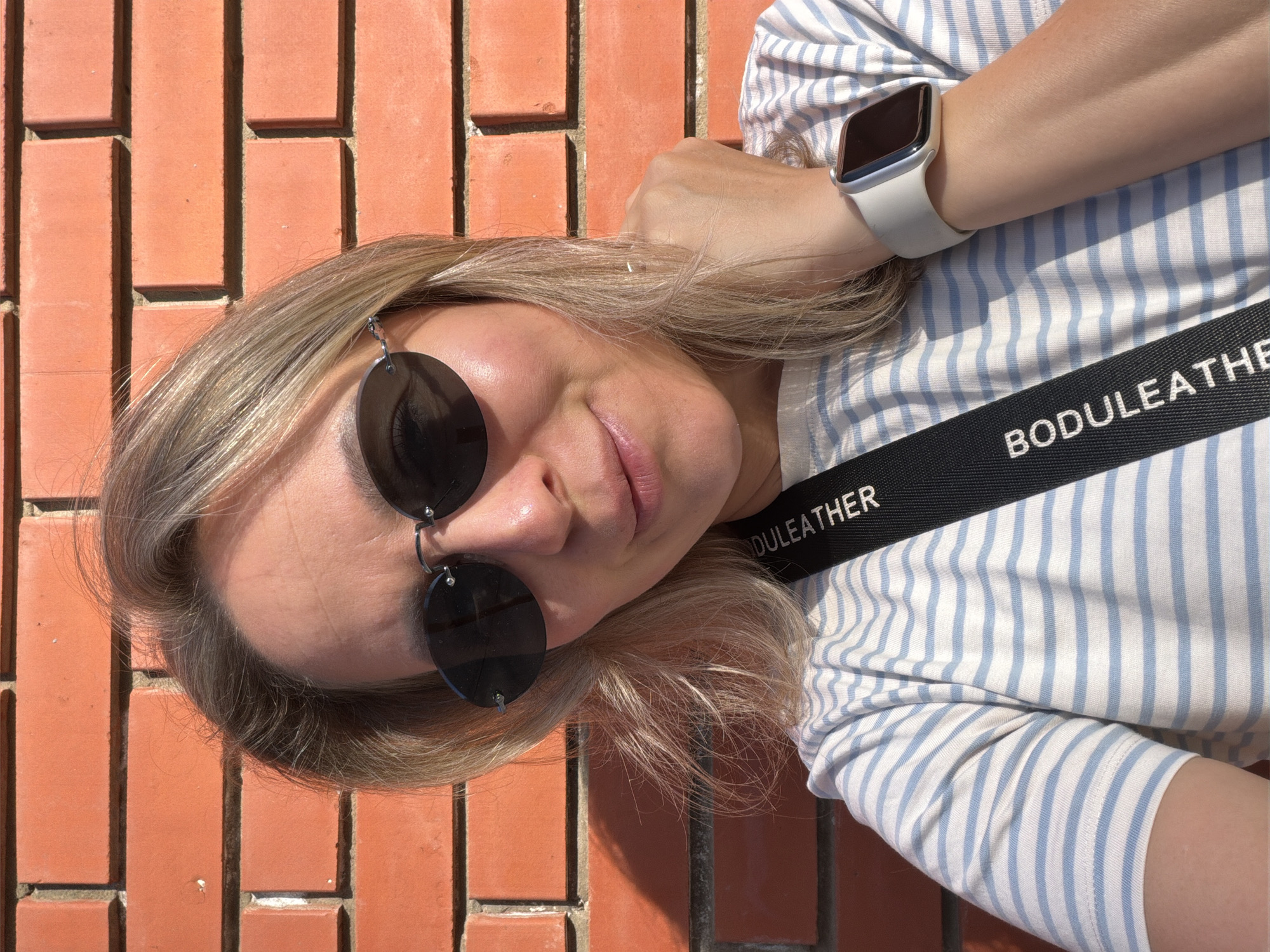 A woman with blonde hair and sunglasses is positioned in front of a brick wall. She is wearing a striped shirt and a white smartwatch. A strap with the text "BODULEATHER" is visible across her chest. The setting appears to be outdoors. There is no indication the image was taken in Chaikovsky, Russia.