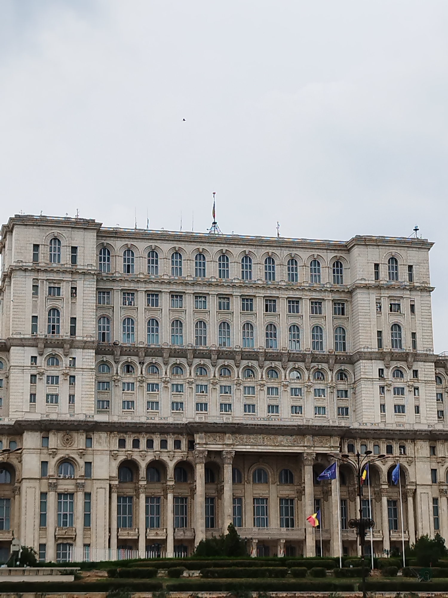 The image shows a large, ornate building in Bucharest, Romania, identified as the Palace of the Parliament. The building is constructed of light-colored stone and features a symmetrical design with multiple rows of windows, many of which are arched. The façade is detailed with columns, balconies, and decorative stonework. Several flags are visible flying in front of the building. The sky is overcast and a single bird is visible in the upper left portion of the image. The foreground shows a landscaped area with manicured bushes and trees, suggesting the building is a significant public monument or government building.