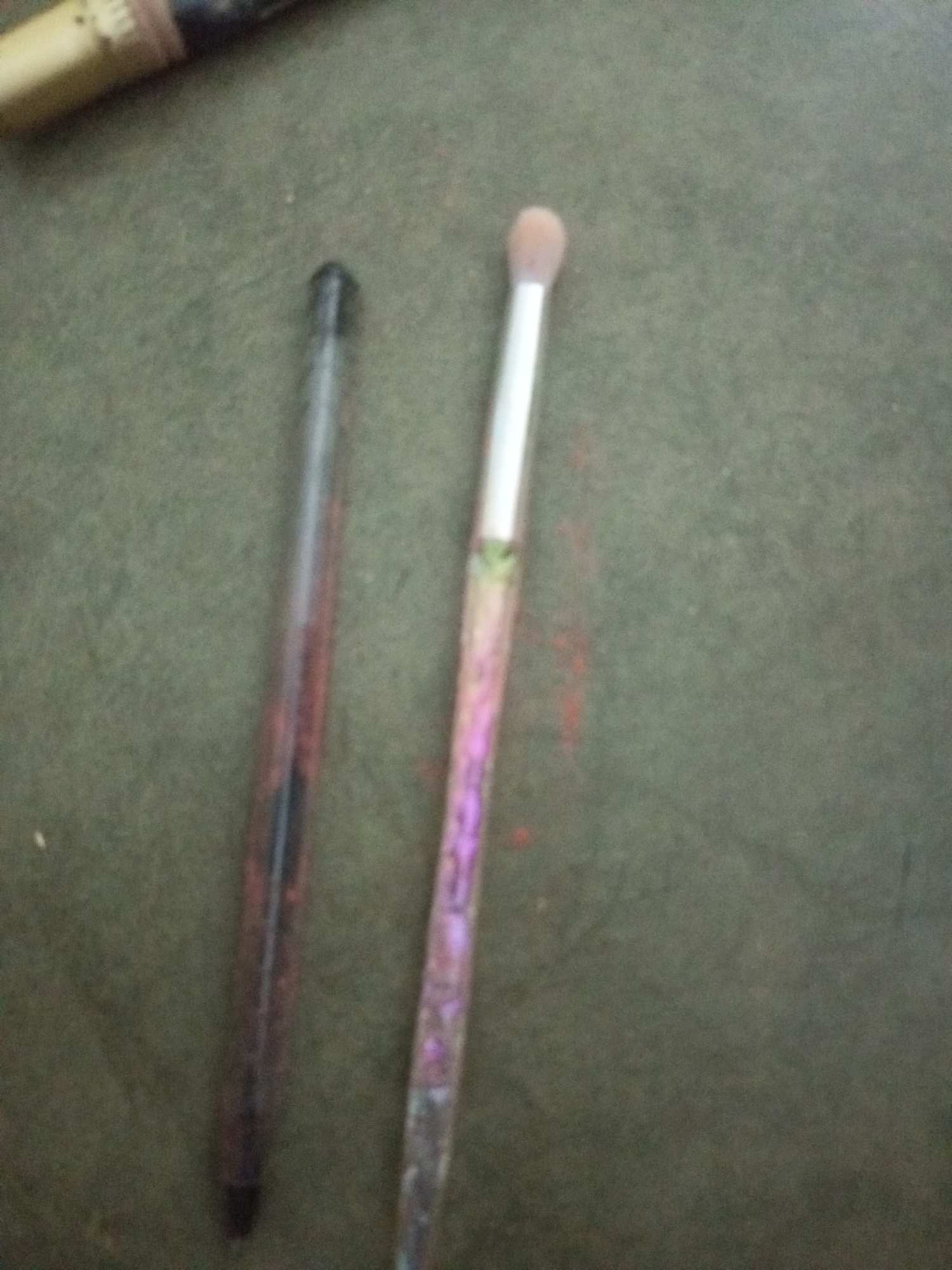 Two makeup applicators are on a dark, textured surface. The applicator on the left is a cylindrical object with a dark body, possibly a pen or a thin makeup brush with a cap on. The applicator on the right is a makeup brush with a silver handle and a fluffy, light-colored brush head. The background appears to be a dark green, rough surface, potentially a textile or a natural material. There are no people or discernible interactions depicted. The location within La Tagua, Venezuela is not identifiable from the visual information provided in the image.