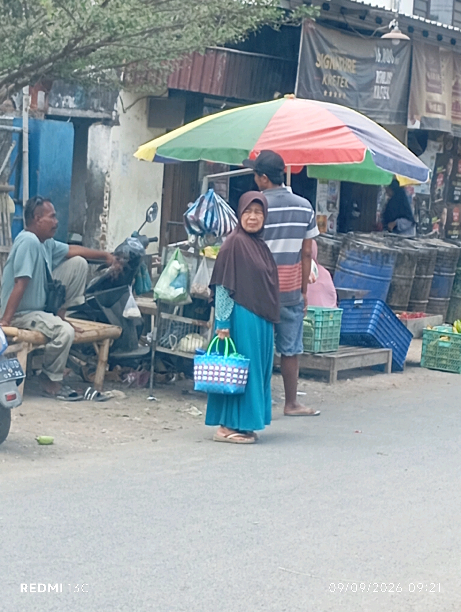 The image depicts an outdoor scene, likely a roadside market, in Wuluhan, Indonesia. In the foreground, a woman wearing a dark brown headscarf and a bright blue dress stands to the right, holding a blue and white woven bag. To her left and slightly behind her, a man in a striped t-shirt and denim shorts stands with his back partially to the camera. Further to the left, a man wearing a light blue t-shirt and light-colored trousers sits on a wooden bench. A motorcycle is parked behind the man on the bench.
The background shows several stalls with various goods, including what appear to be plastic crates containing produce, large blue barrels, and umbrellas. A colorful umbrella, red, yellow, and green, is prominently displayed above the stalls. A banner with text is visible on one of the stalls. The ground is paved with asphalt and some dirt. The overall impression is of a lively, bustling market environment. The timestamp "09/09/2026 09:21" is present in the lower right corner of the image, indicating the time the photograph was taken. The text "REDMI 13C" is visible in the lower left corner, likely indicating the device used to capture the image.