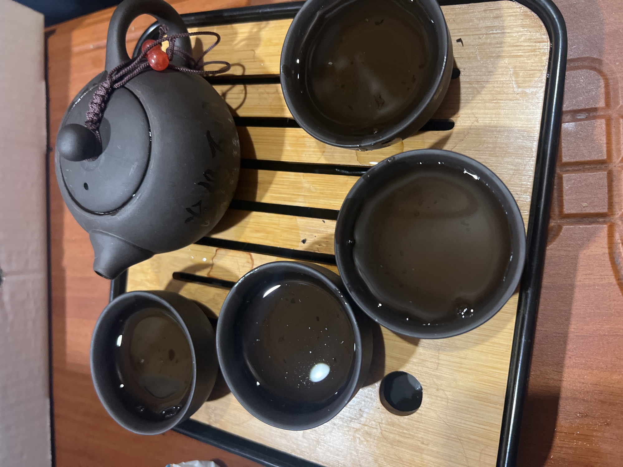A high-angle shot depicts a tea set arranged on a wooden tray with black edging. The tray rests on a wooden surface. A dark gray teapot with a circular handle is positioned on the left. A decorative knot adorns the handle. Three filled tea cups of the same dark gray color are neatly aligned on the right side of the teapot. One additional tea cup is positioned at the bottom left, completing a set of four cups. A small, black disc is placed beside one of the lower right tea cups. The liquid in each tea cup appears to be brown. The specific location is Ильинская Поляна Снт, Russia.