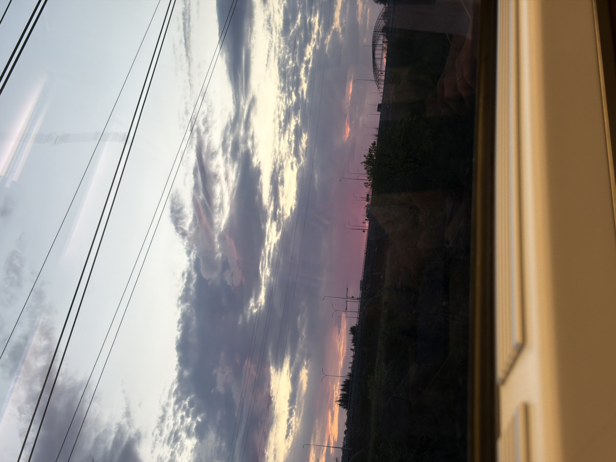 The image shows a view through a window, likely from a moving vehicle, towards a dusky sky with scattered clouds. Power lines are visible across the sky. Below the sky, the landscape includes streetlights and vegetation, seemingly along a road or highway. The overall scene suggests an urban or suburban setting at dusk or dawn. Based on information provided, the location is Japarauli, Georgia. The edge of the window frame is visible on the right side of the image. A reflection is visible in the window. No people are clearly discernible.