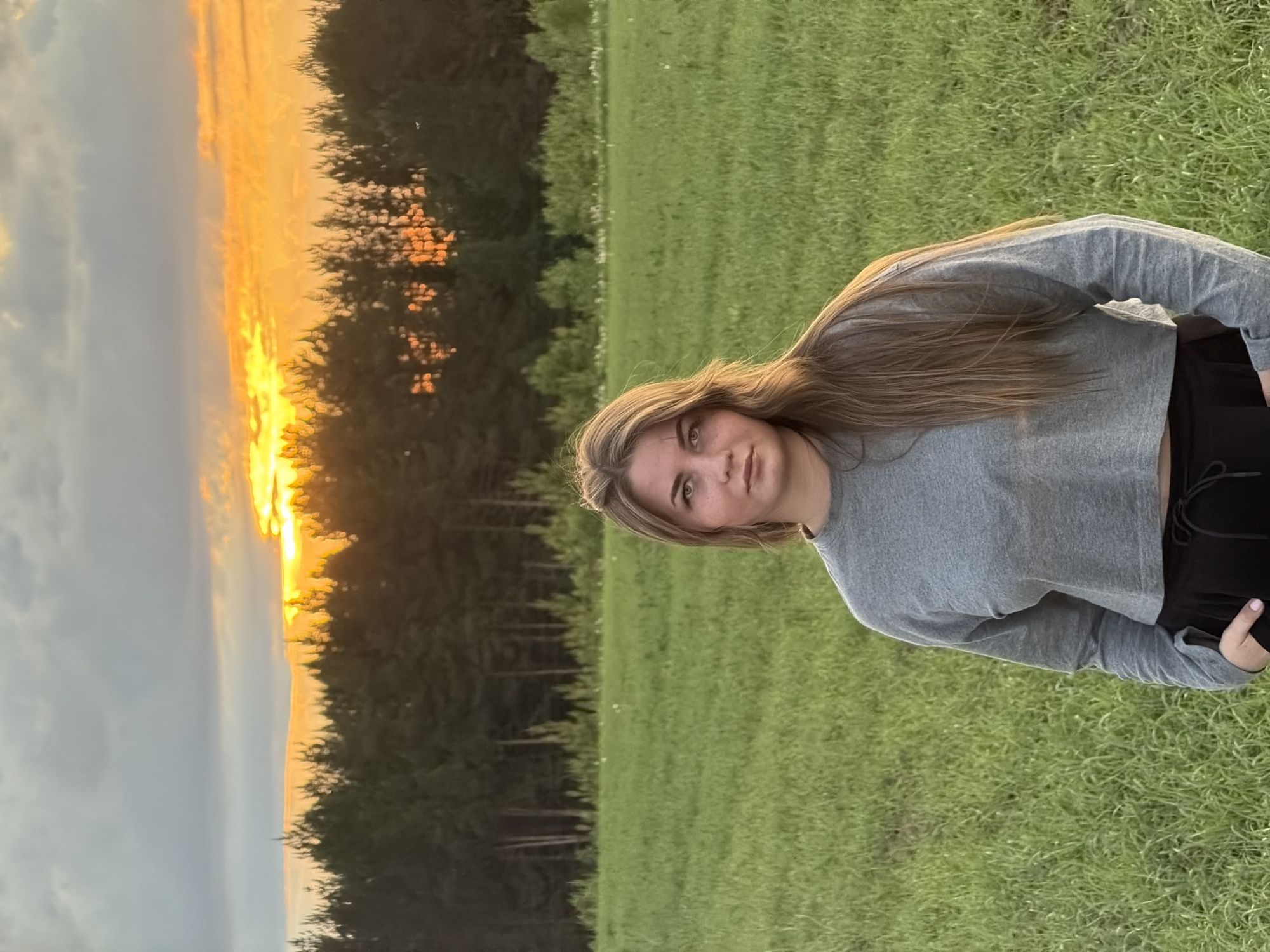 A medium, vertical shot depicts a young, light-skinned woman outdoors at dusk. She is standing in a grassy field with a line of trees and a sunset in the background. The woman is facing slightly away from the camera and has long, light brown hair and a gray, long-sleeved cropped shirt. She is also wearing dark bottoms. The background features a dense line of trees and a sunset with golden and orange hues. The location is likely rural, but the specific city of Фоки, Russia cannot be determined with certainty from the image. There are no visible actions or interactions of interest.