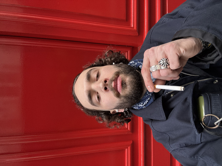 A stylish man with curly hair and a beard poses against a vibrant red door. He wears a navy jacket, a patterned bandana, and multiple rings. He holds a lit cigarette between his fingers. The man has a serious expression, looking directly at the camera. The image focuses on his upper body and face, highlighting his distinctive features and clothing.