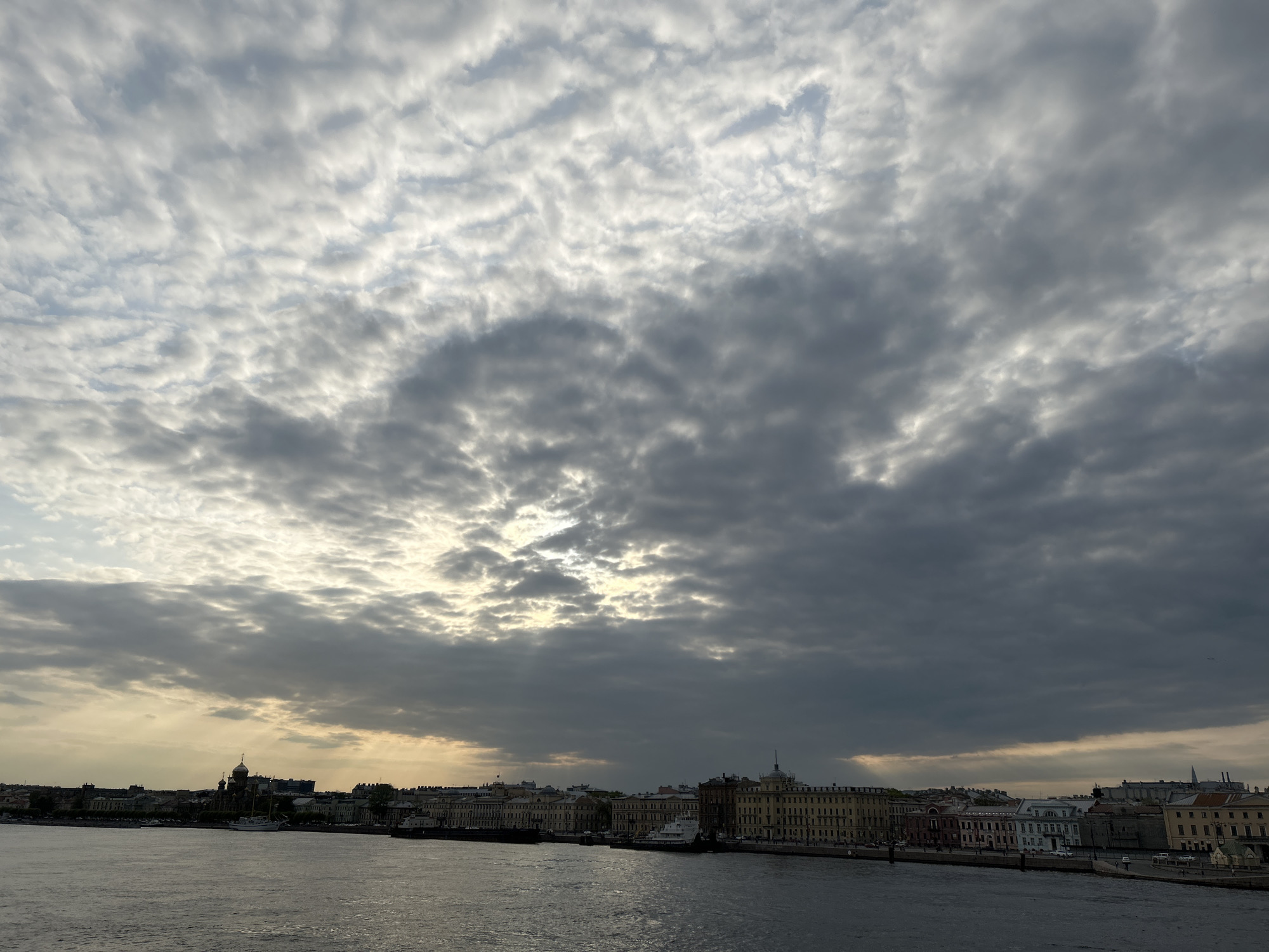 The image depicts a cityscape of Saint Petersburg, Russia, under a cloudy sky. The foreground is dominated by a body of water, likely the Neva River, with ripples visible on its surface. Along the riverbank, a row of buildings stands. The architectural style is classical, with many buildings displaying facades adorned with columns and detailed ornamentation. In the background, the sky is filled with dynamic cloud formations, ranging from light, almost white, to darker gray hues. The overall light suggests either late afternoon or early evening. The atmosphere is overcast.