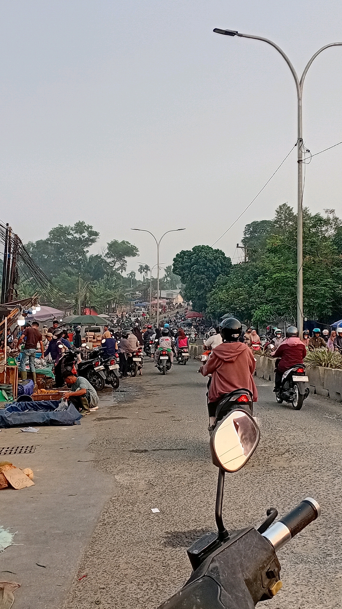 The image depicts a street scene in Jambi, Indonesia, characterized by moderate traffic congestion. The perspective is from the handlebars of a motorcycle, looking forward down a road lined with market stalls on the left and trees and a concrete barrier on the right. Numerous motorcycles, many with riders wearing helmets, occupy the road, suggesting a busy thoroughfare. Some individuals are visible in the market area, engaged in activities such as displaying goods and interacting with potential customers. The sky is hazy, indicating a possibly overcast or polluted atmosphere, and streetlights are visible on tall poles. The overall impression is of a bustling urban environment, likely during daylight hours, with a focus on transportation and commerce.