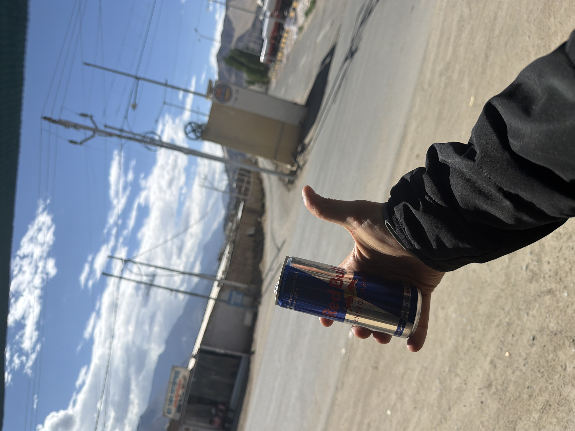 A first-person perspective shot shows a hand holding a Red Bull energy drink can. The hand, with a thumb up, is in the foreground, obscuring the lower portion of the can. The background reveals a street scene in Leh, India, with buildings and mountains under a partly cloudy sky. The can is primarily blue and silver, with the Red Bull logo visible in red. The person is wearing a black jacket. The environment appears to be outdoors, with a paved road and some dirt or gravel on the side. The lighting suggests it is daytime.