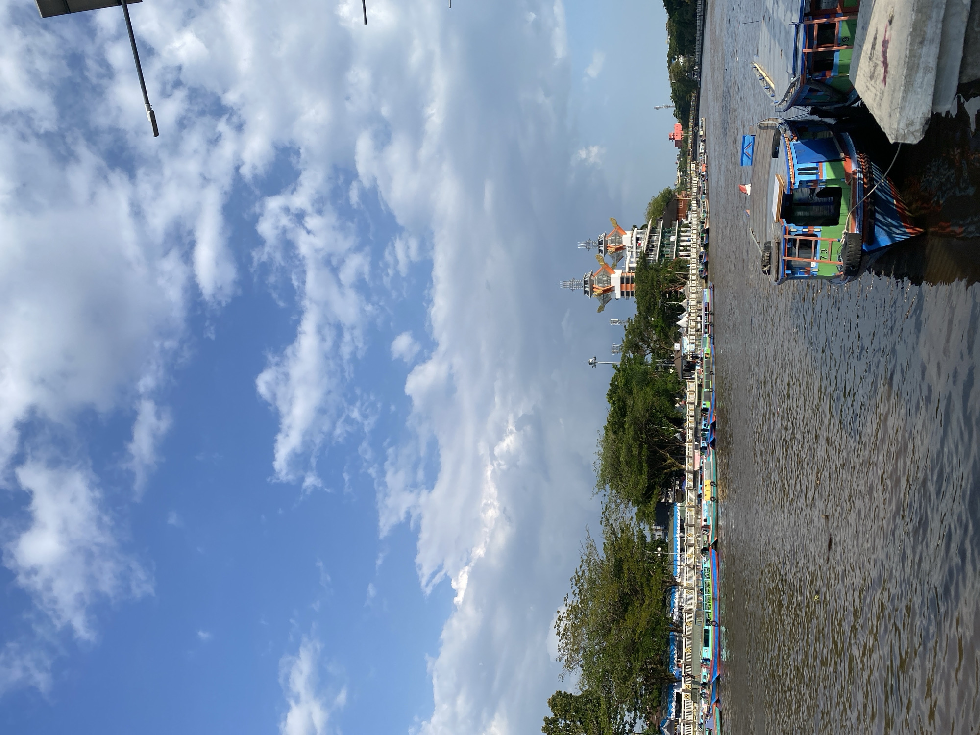 This image captures a view of the Martapura River in Banjarmasin, Indonesia, on a partly cloudy day. In the foreground, two colorful boats are moored near the riverbank, one predominantly blue with green and red accents, displaying the number "73" on its side, and another to its right with green and blue sections. The river itself appears muddy brown with visible ripples and some debris floating on the surface.
Along the opposite bank, a line of trees partially obscures a promenade lined with a white railing and blue awnings. Beyond the trees and promenade, a distinctive building with two prominent, orange-topped structures resembling bird wings or abstract sculptures rises above the surrounding architecture. This building is a notable landmark in the city. Further in the background, a bridge can be seen spanning the river, and more buildings and trees line the distant riverbanks. The sky above is a bright blue, dotted with white, fluffy clouds. The overall scene suggests a lively urban waterway with a blend of traditional and modern elements.
