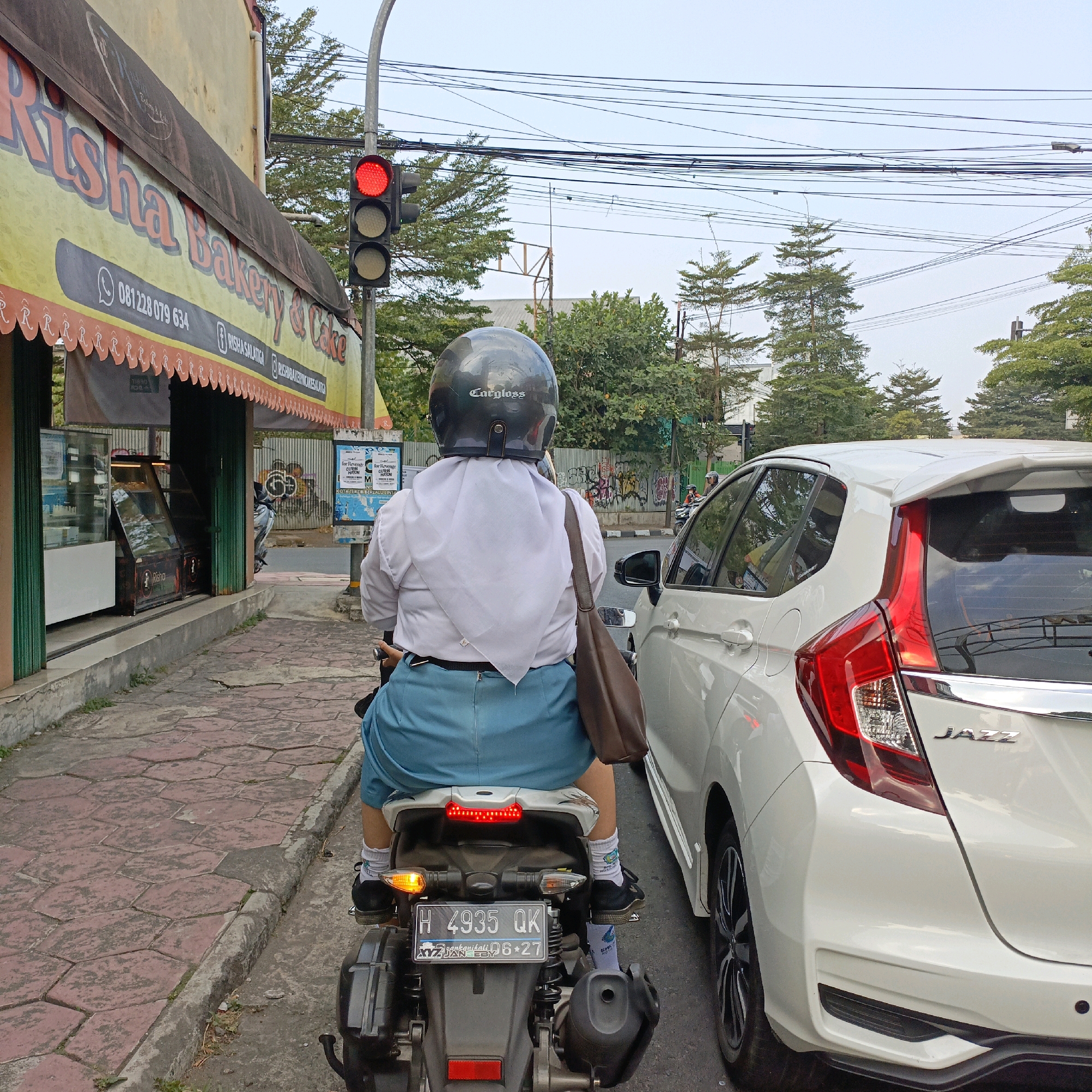 The image captures a street scene in Salatiga, Indonesia, likely near a bakery given the signage for "Risha Bakery & Cake." The perspective is from behind a person riding a scooter, who is wearing a white shirt, blue shorts, and a dark helmet with "Catgloss" branding. They are stopped at a traffic light, which is red. To the right of the scooter is a white car, identified as a Honda Jazz by its badging. The background shows trees, power lines, and some graffiti on a wall. The pavement is made of hexagonal red tiles. The overall impression is a typical urban traffic situation.