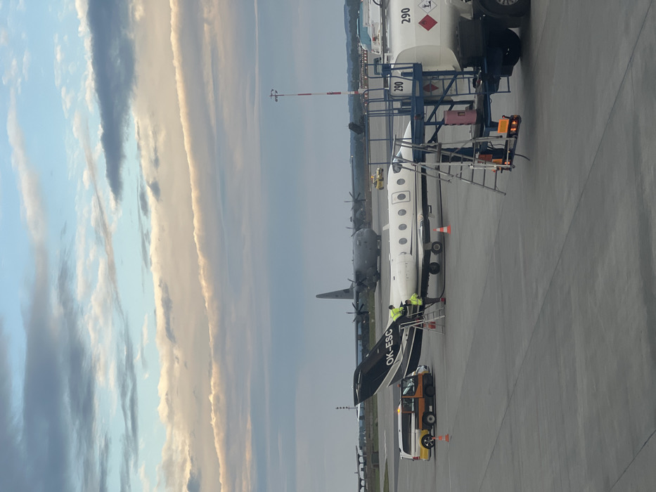 The image shows a private jet being refueled at the Rzeszow-Jasionka Airport (RZE) in Jasionka, Poland. The coordinates 50.114, 22.026 are consistent with the airport’s location. The jet is a white and black Cessna Citation CJ4 with the registration number OK-ESC. There is a fuel truck parked next to the jet, connected to it via a hose. Two people in high-visibility vests are interacting with the jet. In the background, a larger aircraft, likely a military C-130 Hercules, is parked and facing the camera. The image was taken on a cloudy day, with a clear view of the runway and tarmac. The ground crew appears to be professionally carrying out the refuelling operations.