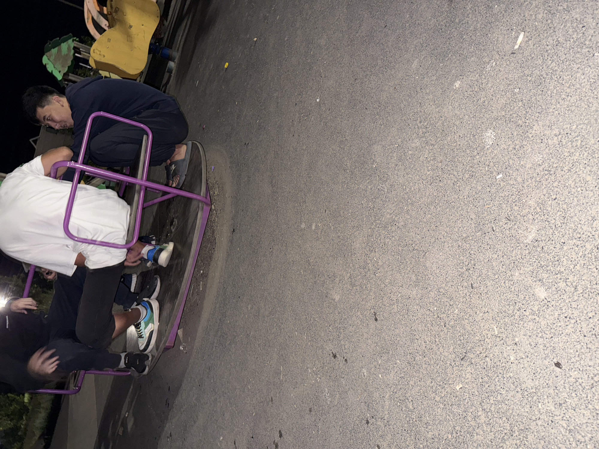 The image shows a group of young men sitting on a purple merry-go-round in a park at night. The ground is paved with asphalt. The men are dressed casually in dark clothing. One man is wearing a white t-shirt and dark pants, another is in a dark hoodie and dark pants, and a third is in a black sweatshirt and black pants. They are all sitting on the platform of the merry-go-round, with their legs extended. The merry-go-round is stationary. In the background, there is a yellow playground structure. The lighting suggests it is nighttime. The location is identified as Zaşağan, Kazakhstan.