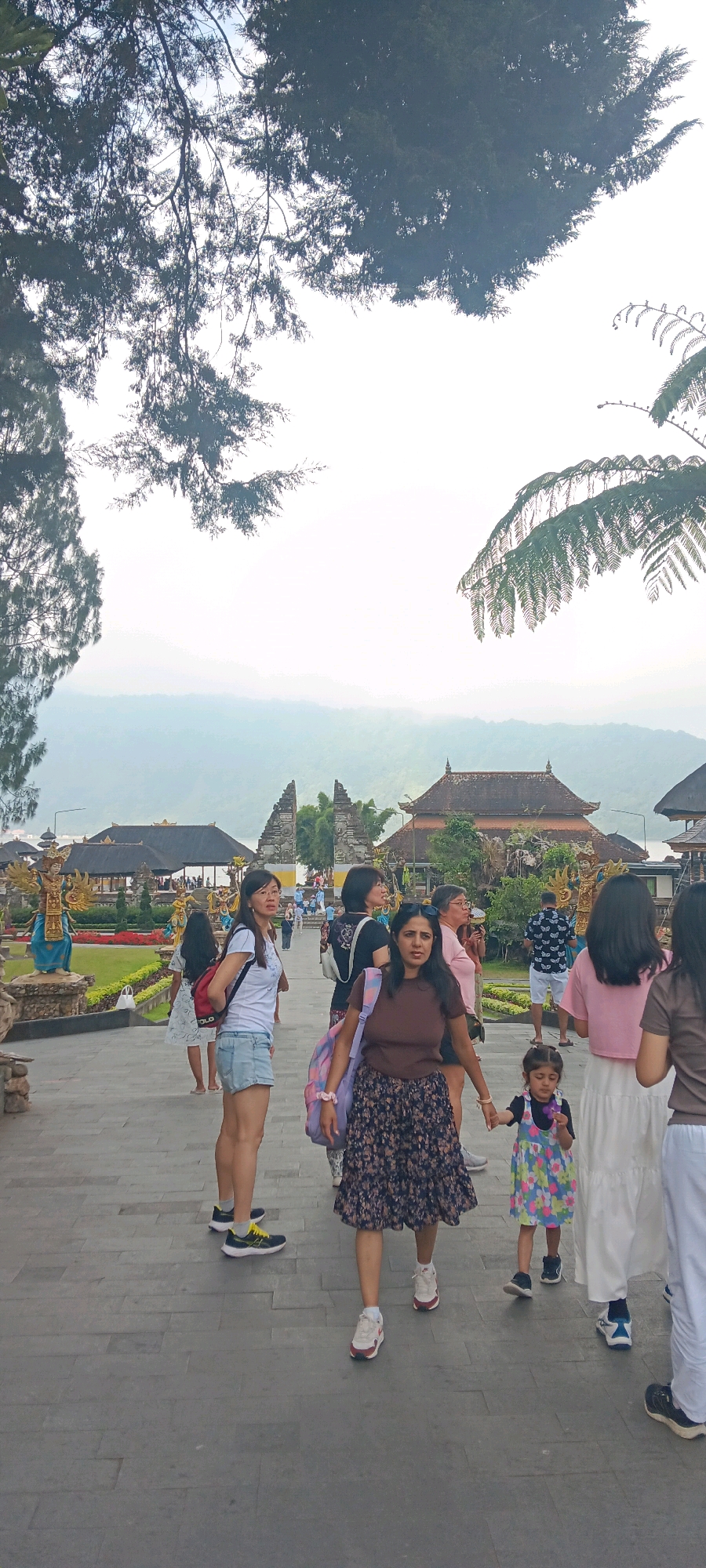 In Baturiti, Indonesia, a group of people are walking along a paved pathway in what appears to be a temple complex. The pathway is made of large, grey rectangular stones. On the left side of the path, there are manicured gardens with colorful flowers and a statue of a figure in a blue and gold garment. Further back, traditional Balinese architecture is visible, including tiered roofs and ornate gates. In the background, mist obscures rolling hills and mountains.
The people in the foreground include several women and a young child. One woman in the center is wearing a brown t-shirt and a floral print skirt. She is holding the hand of a young girl in a colorful floral dress. To her left, a woman in a white t-shirt and denim shorts is walking with a backpack. Another woman in a pink t-shirt and white skirt is walking to the right. In the middle ground, other individuals are seen walking and standing. The overall atmosphere is one of a casual outing, with people dressed in light clothing suitable for warm weather. The sky is overcast, creating a diffused light.