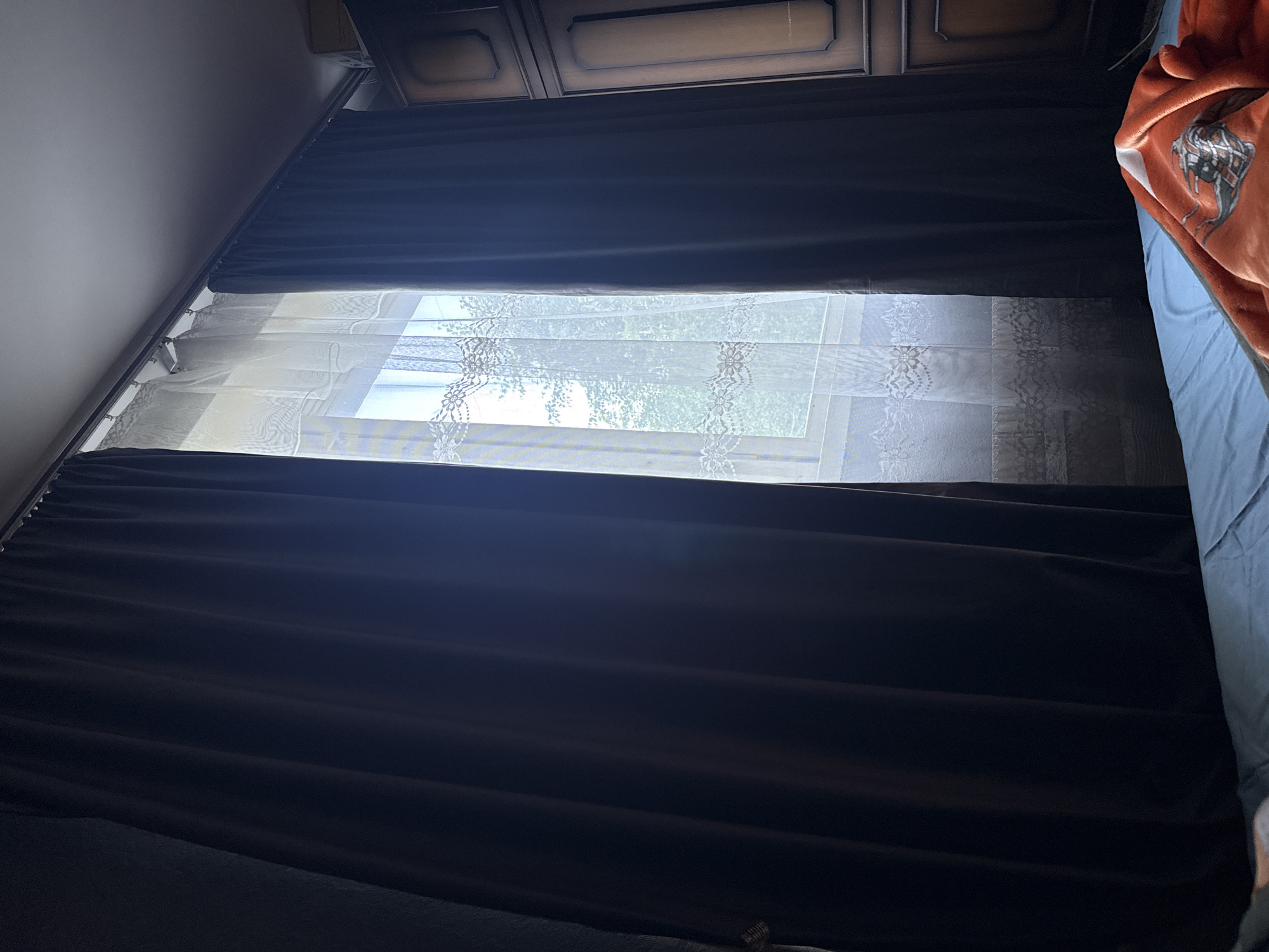 An indoor scene depicts a window covered by dark brown curtains and a sheer white lace curtain. Sunlight streams through the window, illuminating the lace pattern. To the right of the window is a wooden wardrobe with carved details. In the foreground, a bed with blue sheets and an orange blanket is visible. The scene appears to be in a residential setting. The image does not provide enough information to determine the specific location in Moscow, Russia.