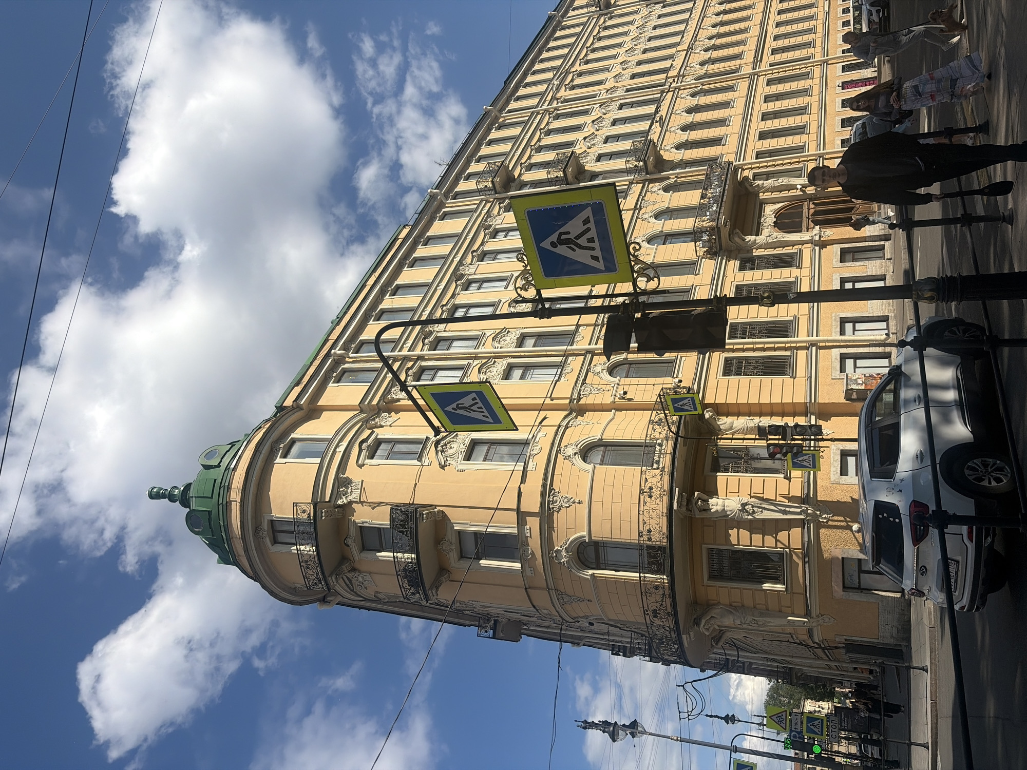 The image shows a building in Saint Petersburg, Russia, with a distinctive curved facade and a green dome. The building is yellow and features classical architectural elements, including balconies with ornate railings and decorative statues supporting the upper floors. Several pedestrian crossing signs are visible, indicating a street with traffic. A white car is parked on the street, and several people are walking on the sidewalk. The sky is blue with scattered white clouds, and the sun is shining, casting shadows. The street appears to be paved with asphalt. The building's design suggests it is in a historic district of the city.