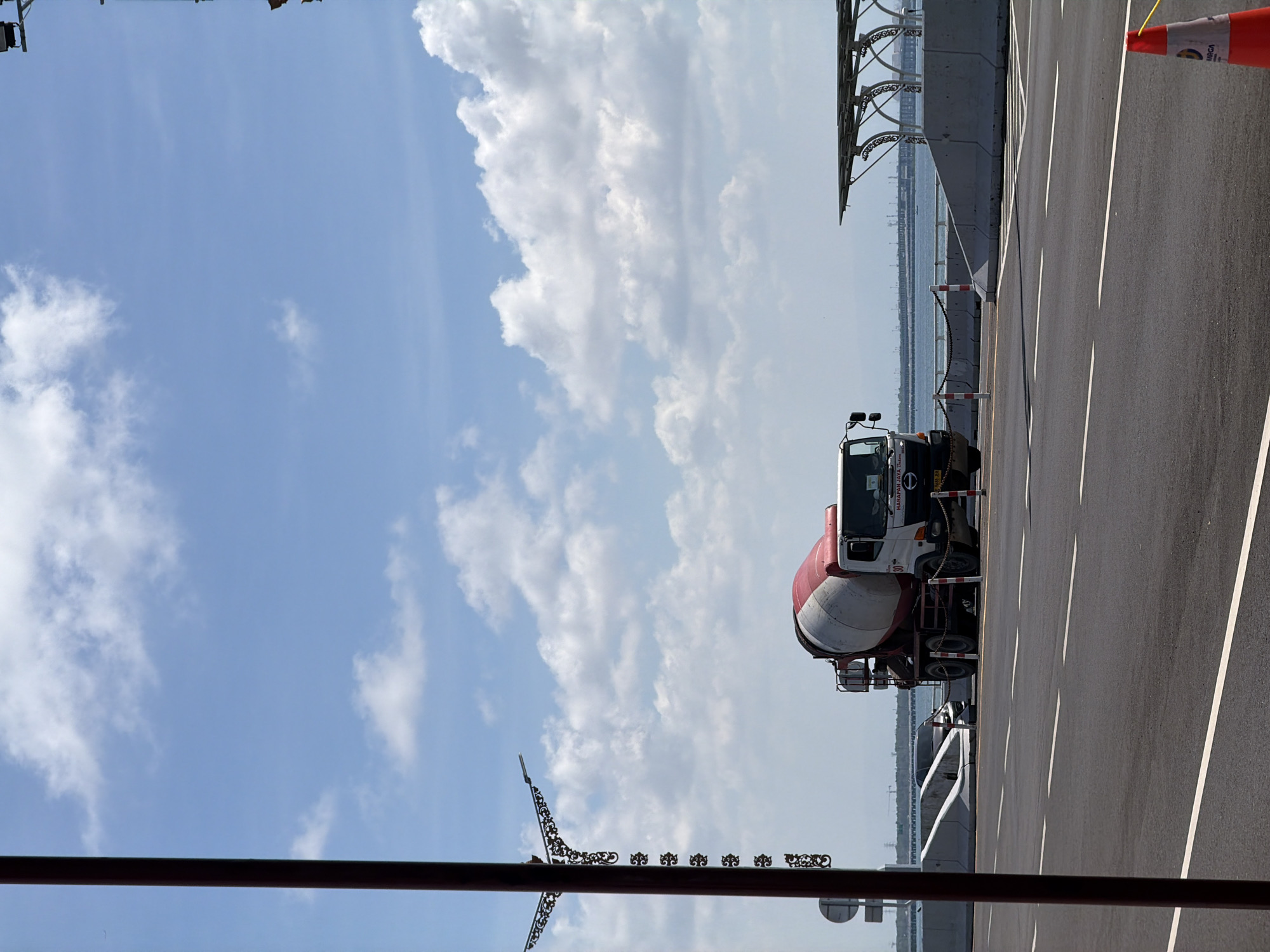 A cement mixer truck is parked on the side of a road, likely under construction or recently paved, with a view of the ocean and a bridge or elevated structure in the background. The truck is white with a red drum. A lamppost with decorative elements stands to the left of the frame. The sky is blue with scattered clouds. The scene appears to be in a coastal area of Kuta Selatan, Indonesia, possibly near a new development or infrastructure project given the presence of construction materials and barriers. There are no people visible in the image.